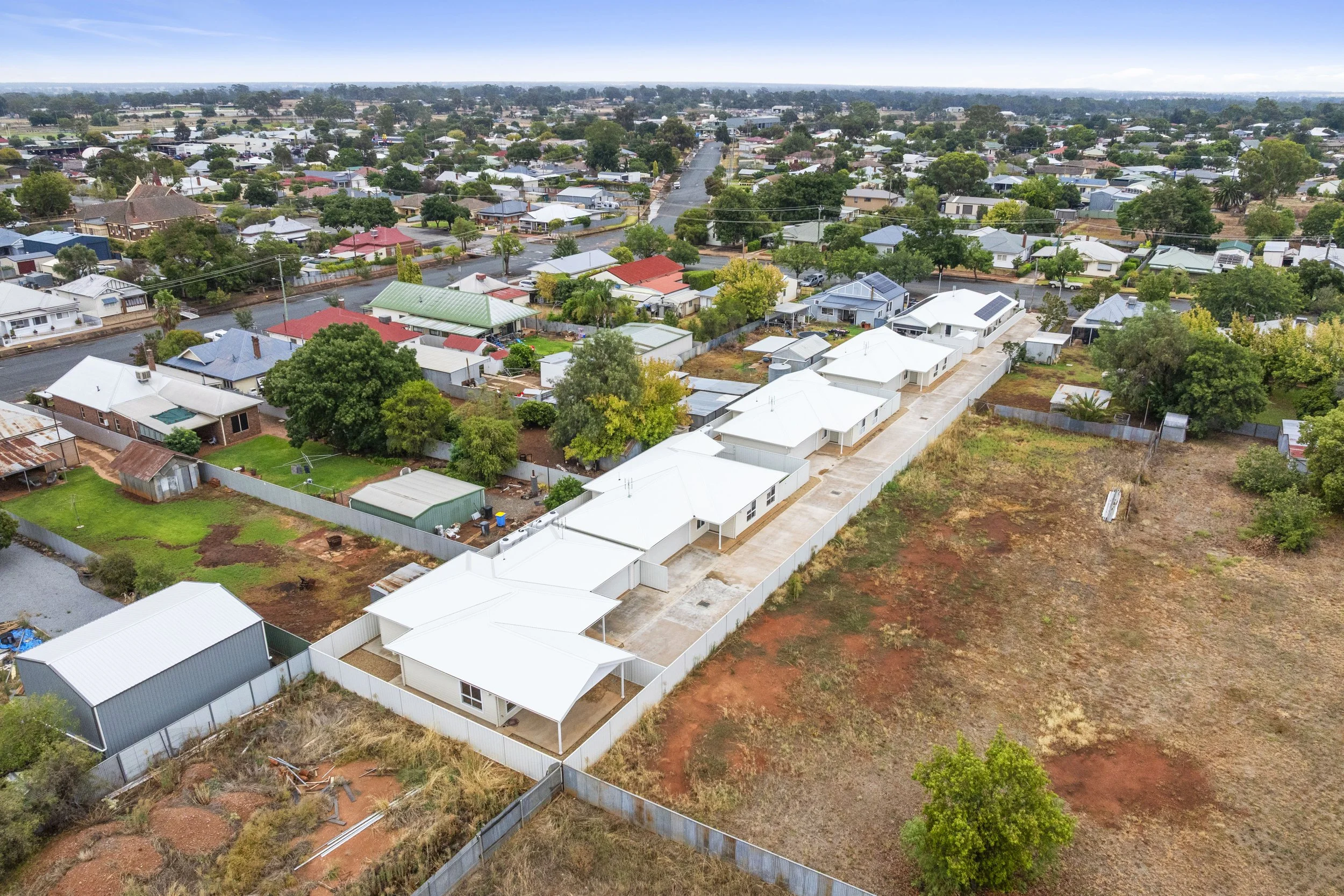 Aerial view of a residential neighborhood with houses, trees, and streets. Several houses have white roofs and are lined up along a concrete driveway. There are open lots and fenced yards with some greenery, alongside a flat landscape with sparse veg