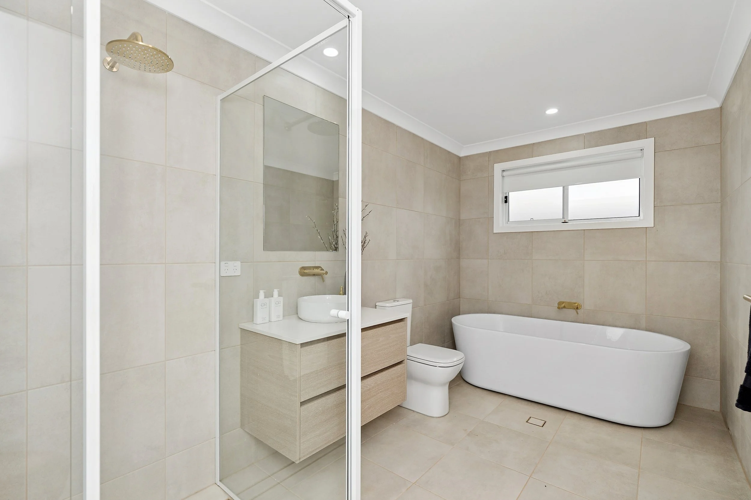 Modern bathroom with a separate shower stall, a white bathtub, a toilet, and a vanity with a round sink. Beige tiled walls and floor, a small window, and minimal decor.