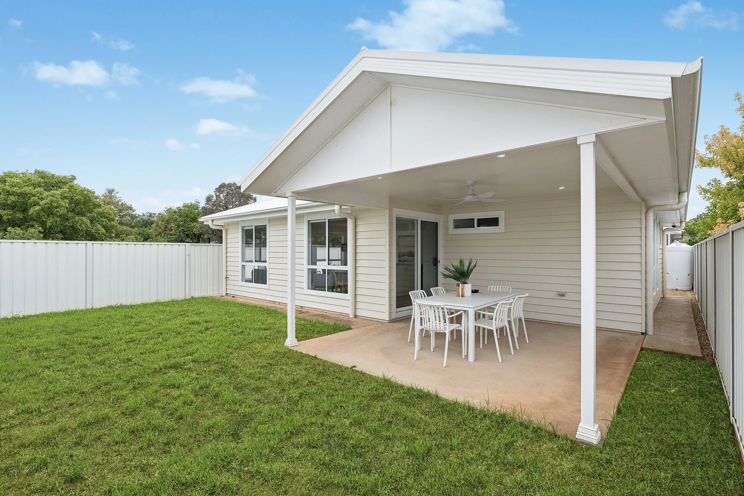 Backyard with a covered patio, white house with large windows, white fence, green grass, and a table with chairs.