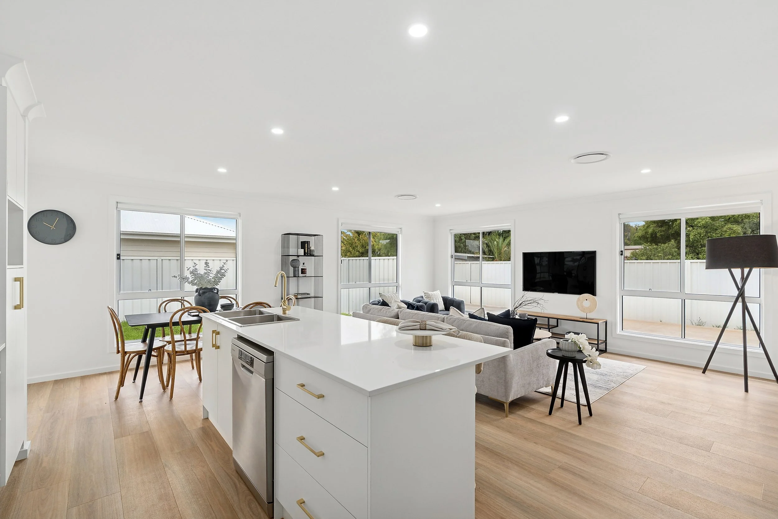 Open-concept living room and kitchen with white walls, light wood flooring, large windows, a white island with gold handles, a dining table with four chairs, a black clock, a TV on the wall, a floor lamp, and a seating area with sofas and a coffee ta