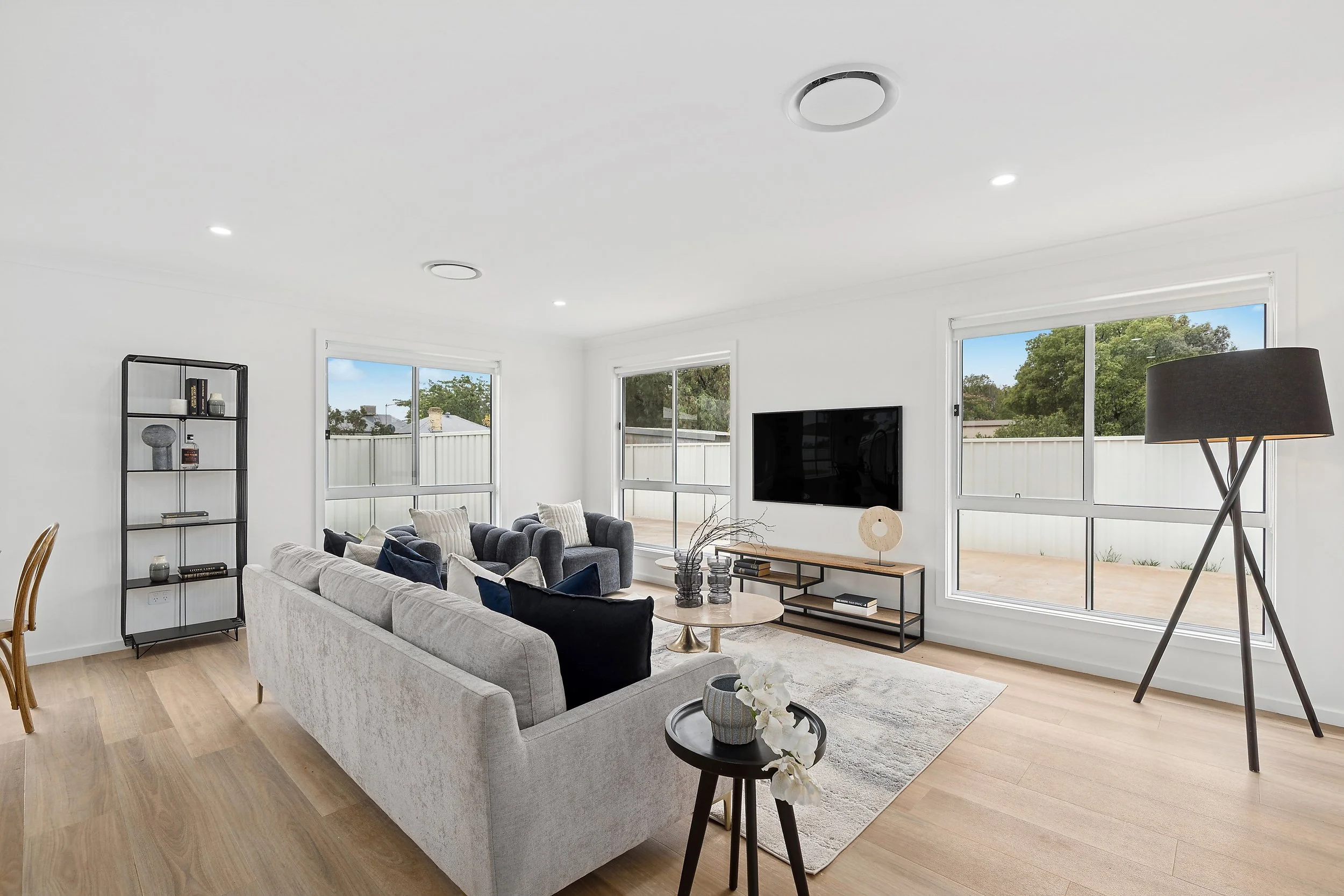 Bright living room with white walls, large windows, light wood flooring, and contemporary furniture including a gray sofa, matching armchairs, a black bookcase, a flat-screen TV, a black tripod floor lamp, and decorative items on tables and shelves.
