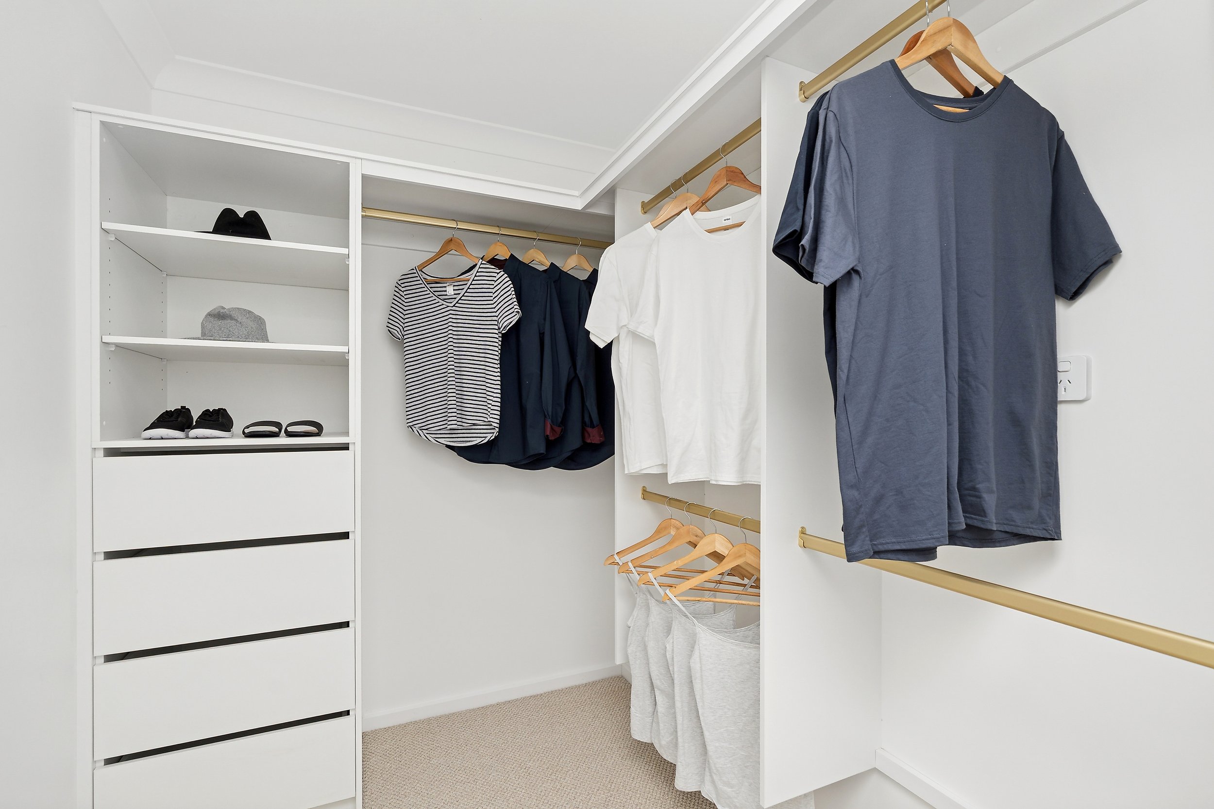 Walk-in closet with organized clothing, shoes, and accessories. White shelves and hanging rods hold shirts, t-shirts, and pants.
