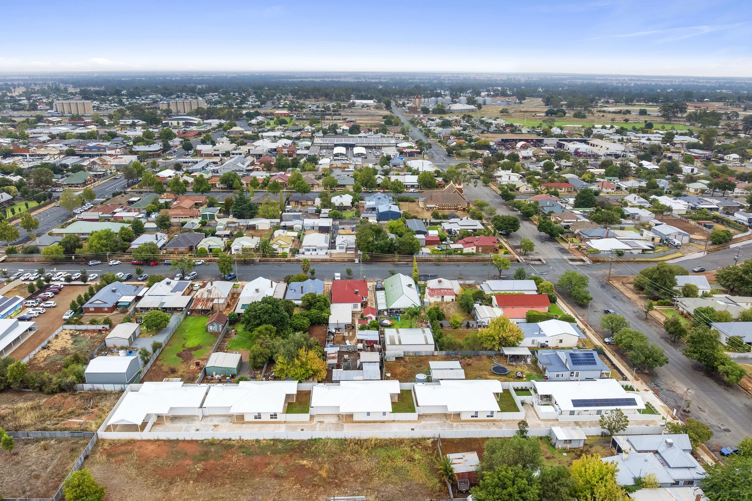 Aerial view of a suburban neighborhood with houses, streets, trees, and open land under a clear sky.