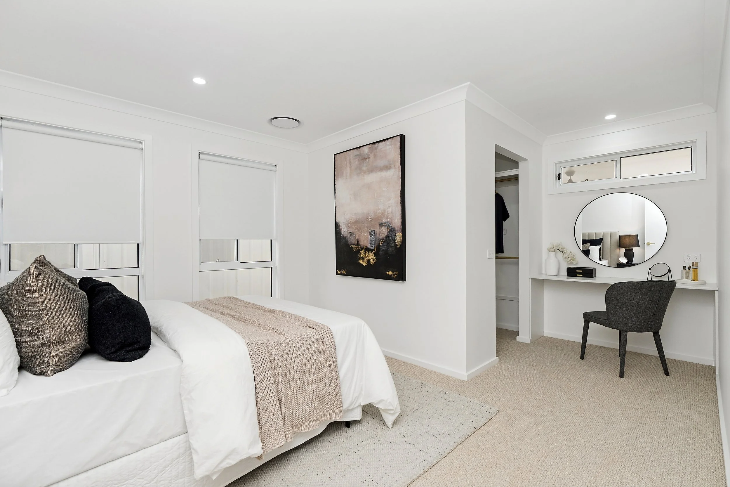 Modern bedroom with white walls, a bed with white linens and decorative pillows, a sitting area with a vanity mirror, and artwork on the wall.