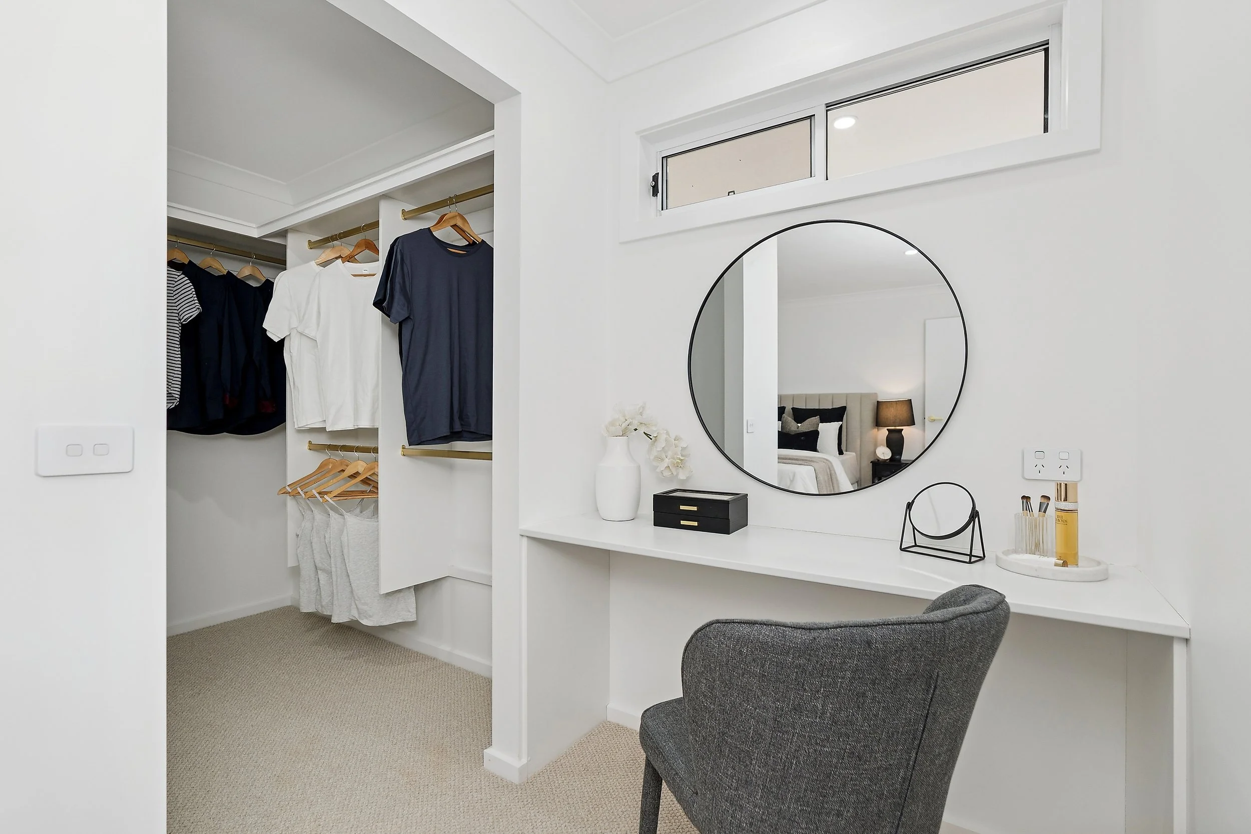 A walk-in closet with hanging clothes and a vanity area with a round mirror, a black jewelry stand, and a small tray with bottles, in a bedroom with neutral tones.
