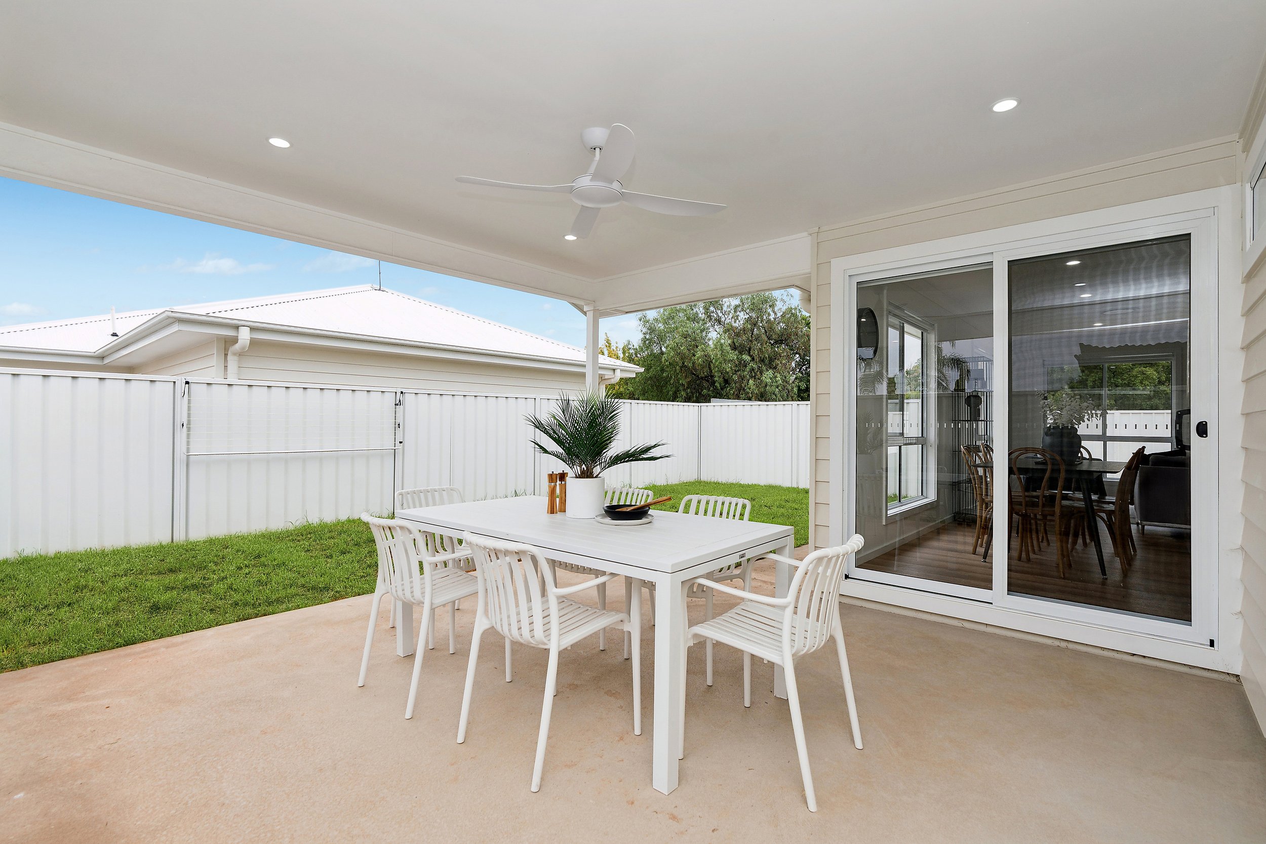 Outdoor patio with a white dining table and chairs, decorated with a potted plant and tableware, enclosed by a white fence and attached to a house with sliding glass doors, leading to an interior dining area.