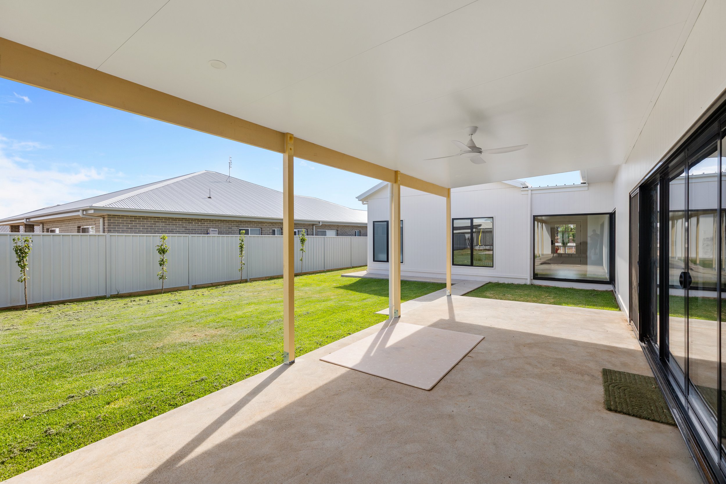 Covered patio area with a ceiling fan, overlooking a grassy backyard with a white fence and a modern house with large windows.