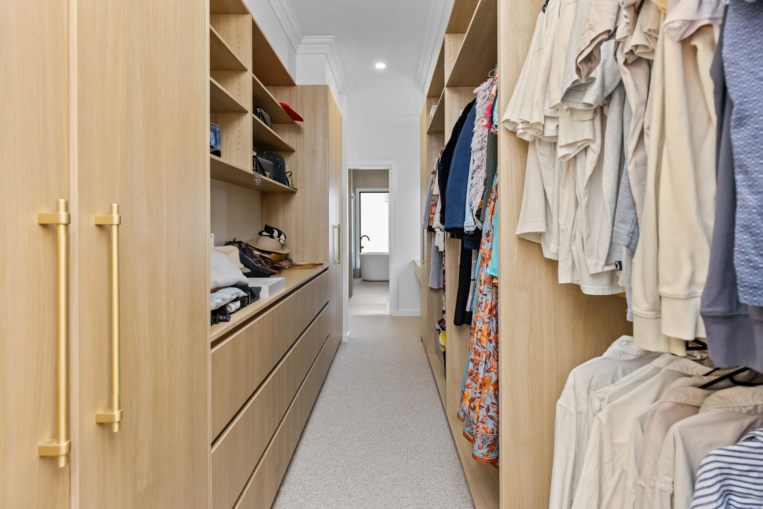 A walk-in closet with wooden cabinets and open shelving filled with clothes, hats, and accessories, leading to a bathroom with a bathtub visible through an open doorway.