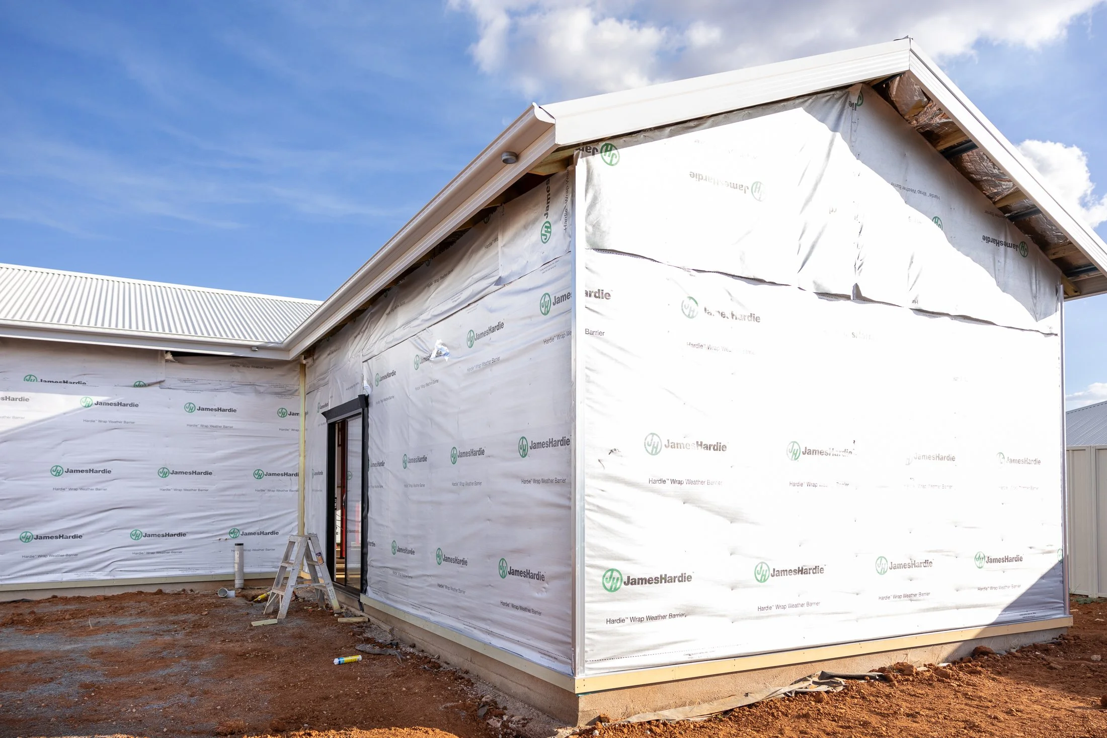 Under construction house wrapped in weather barrier, with a metal roof and dirt ground in front.