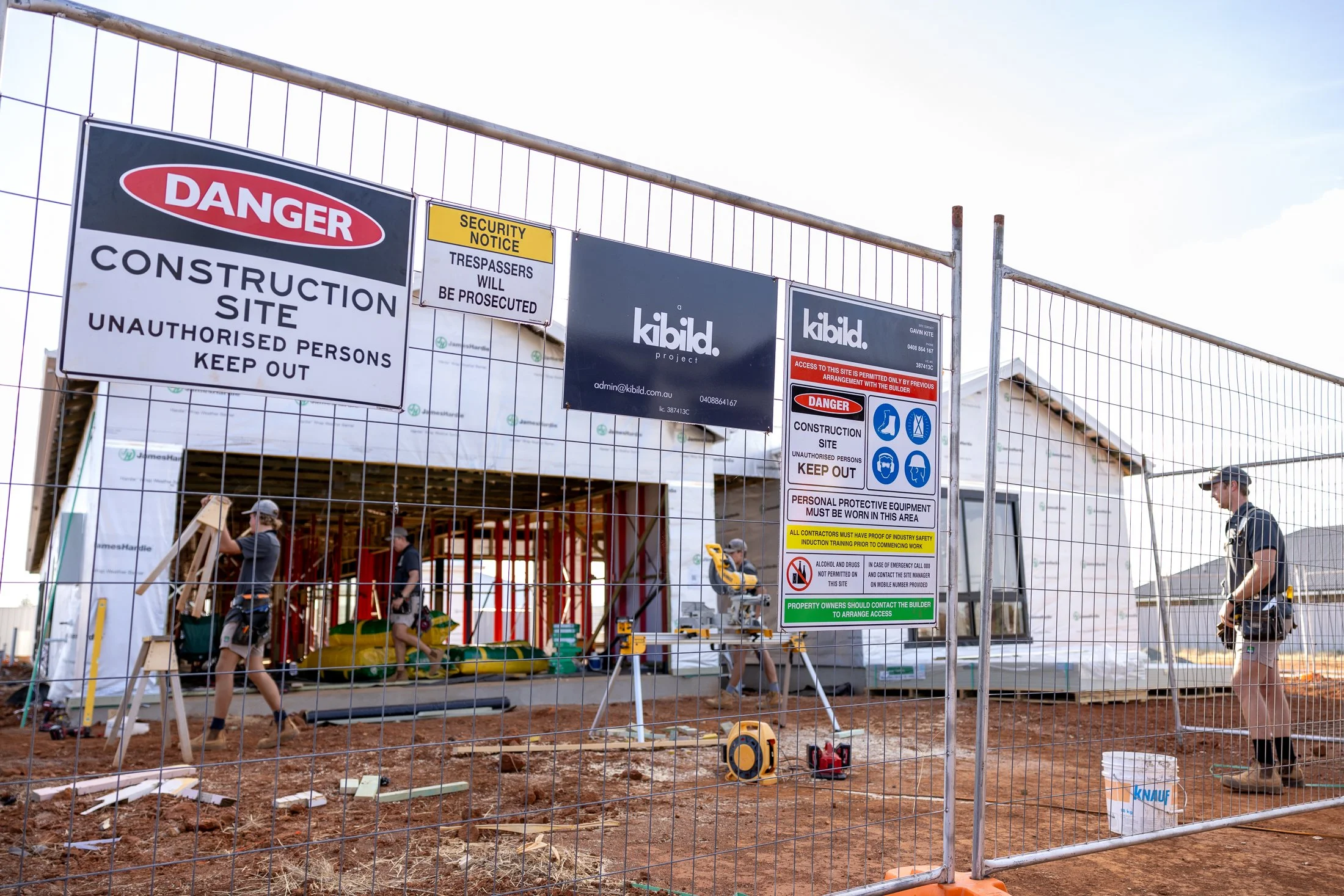 Construction site behind a fence with multiple warning signs, workers wearing safety gear, and construction materials visible.