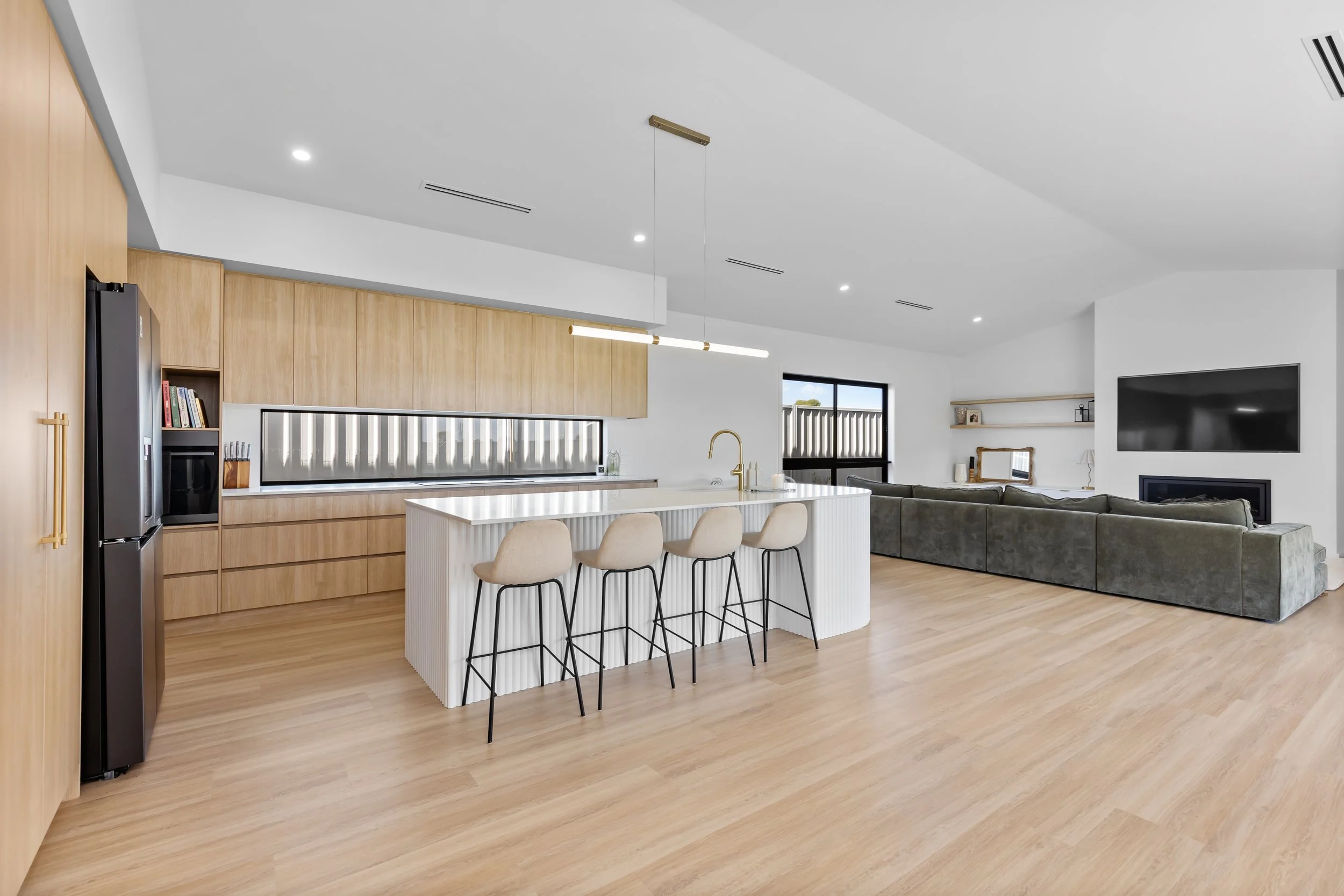 Modern open-concept living room and kitchen with wooden cabinetry, white island with four beige barstools, gray sectional sofa, wall-mounted TV, and minimal decor.