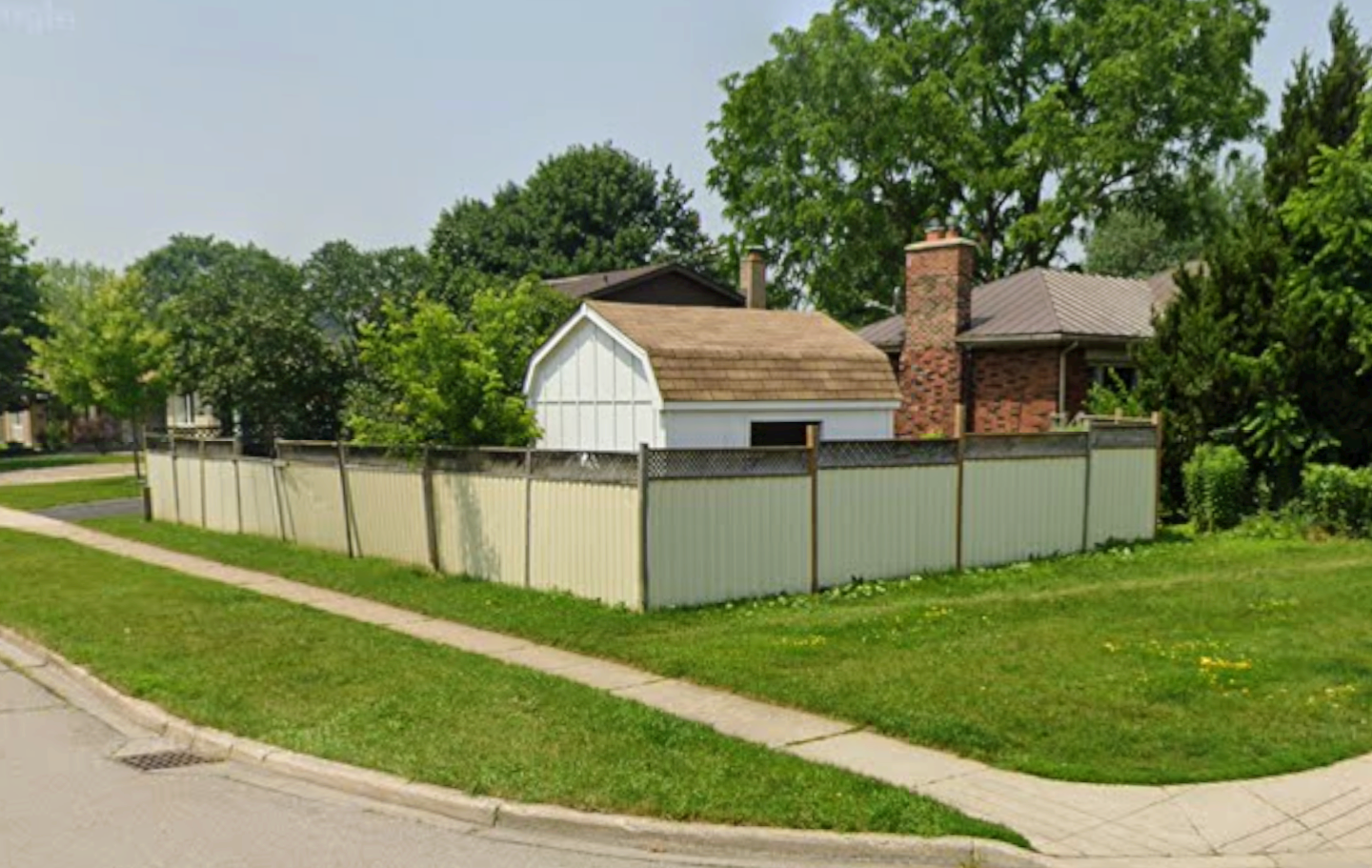 Residential backyard enclosed by a white fence, with a small white shed, red brick chimney, and large green trees surrounding the yard, next to a sidewalk and grassy lawn.