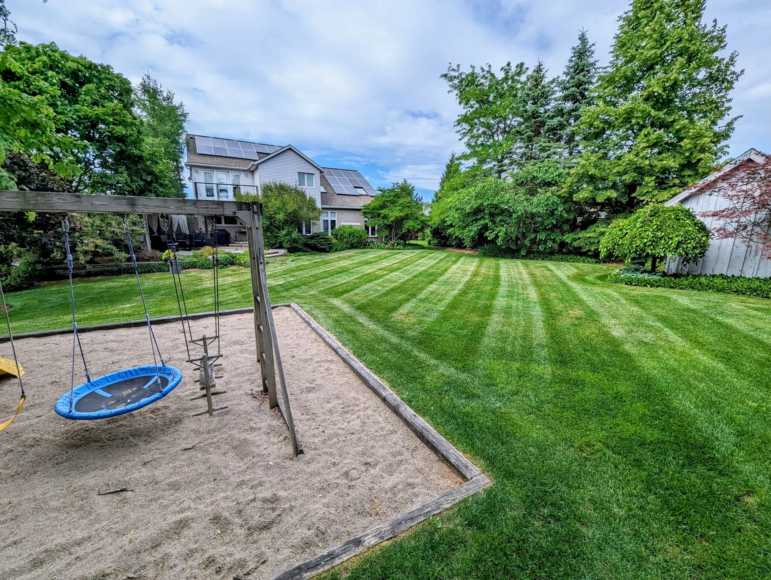 A backyard with a sandbox and swings on the left, a newly mowed green lawn in the center, and trees and houses in the background under a partly cloudy sky.