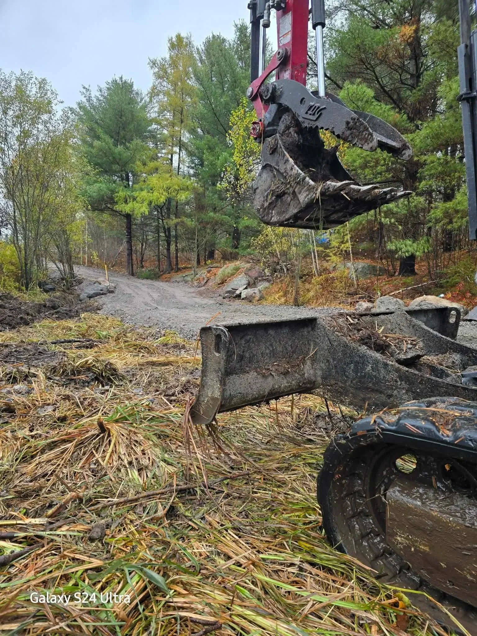 Close-up of a construction excavator bucket and track on a muddy, gravelly forested area with a dirt road in the background.