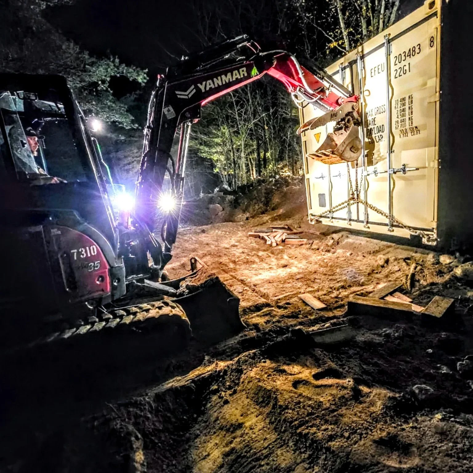 Nighttime scene showing a large excavator moving dirt near a fallen shipping container with a chain and chain hoist attached.