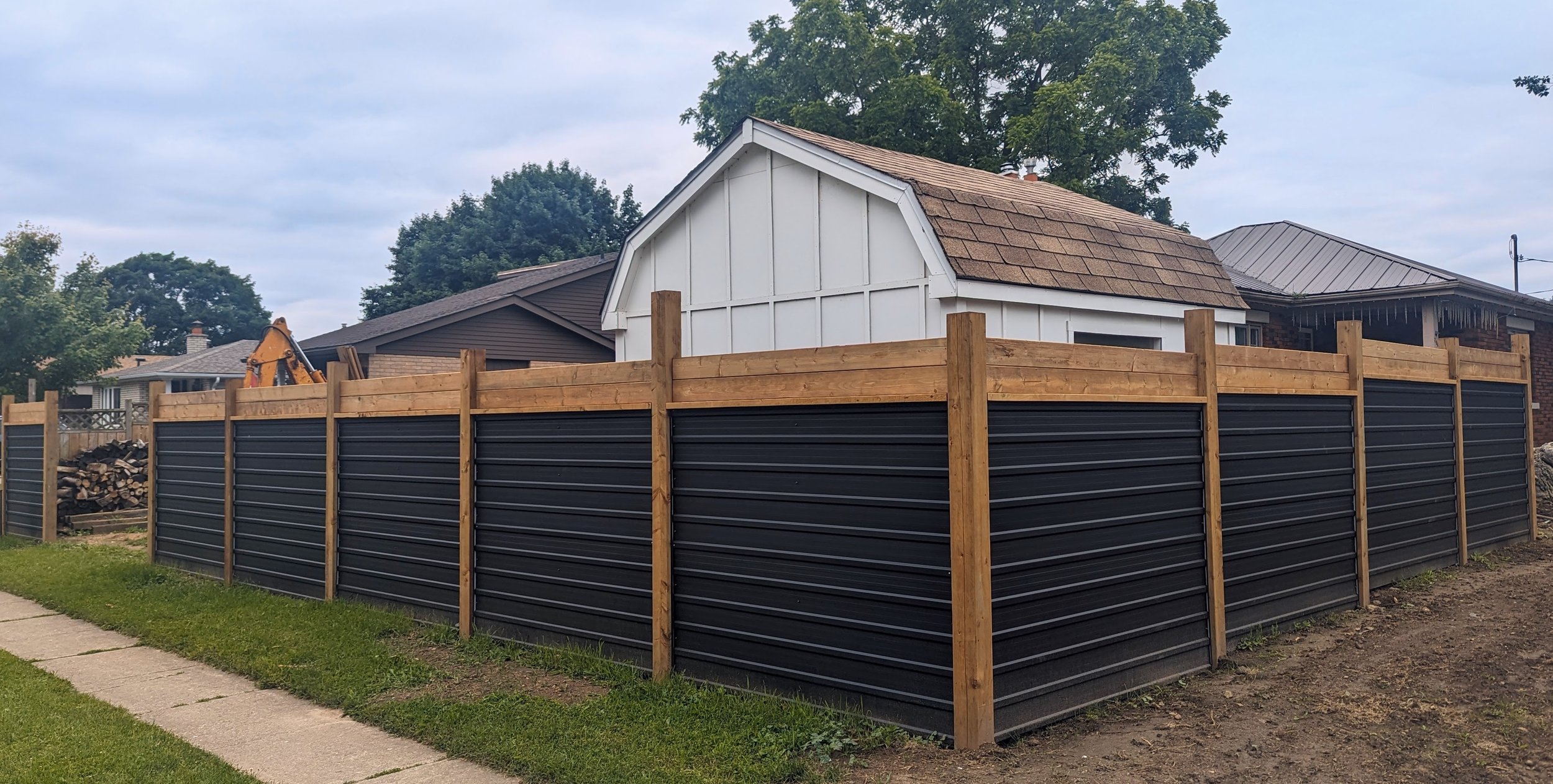 A backyard with a wooden and metal fence, a small shed with a shingled roof, and some trees and houses in the background.