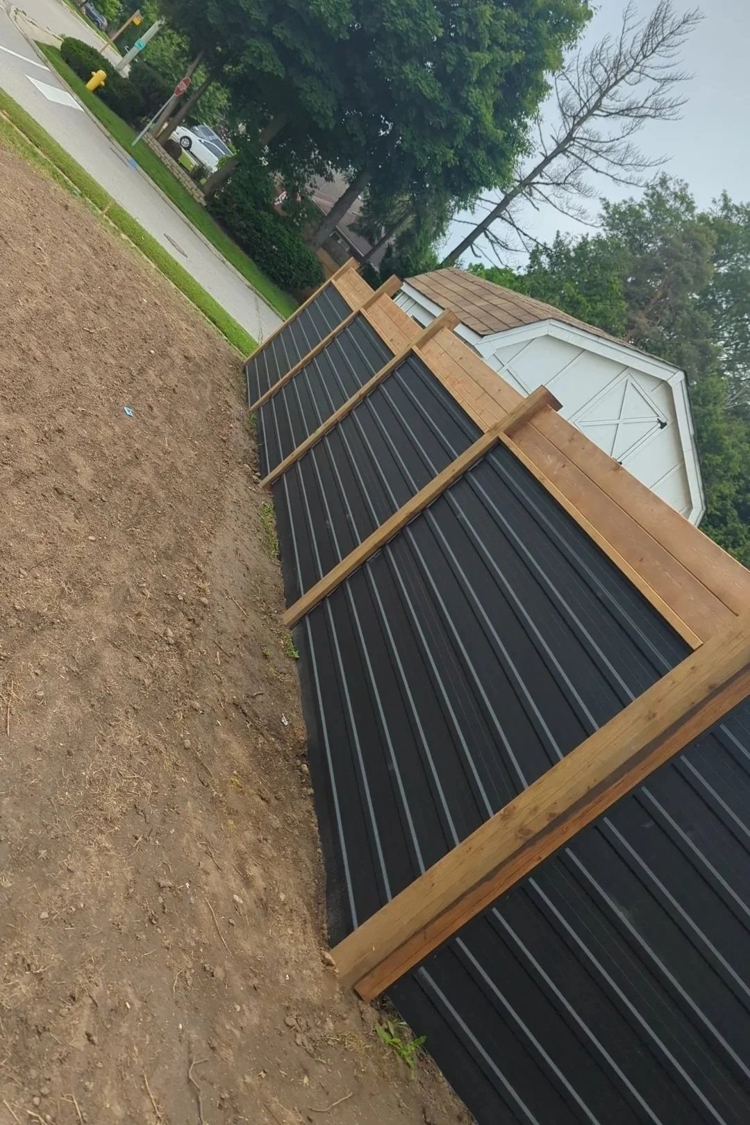 A new black and gray horizontal striped fence with wooden support beams and trim, installed along a dirt patch in a residential yard.