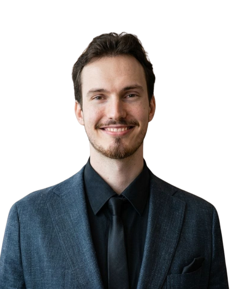 Portrait of a young man with brown hair smiling, wearing a navy blue suit and black shirt, against a white background.