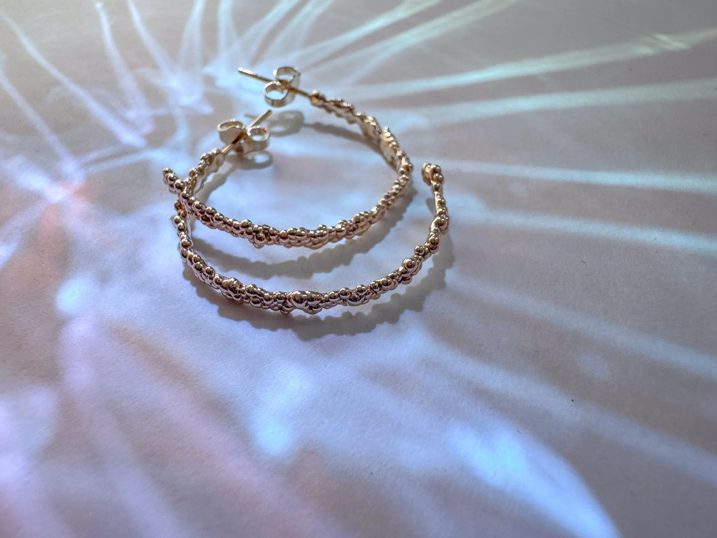 Close-up of 14ct gold Eos hoop earrings with textured bubble design, illuminated by light creating colorful reflections and shadow patterns on a white surface.