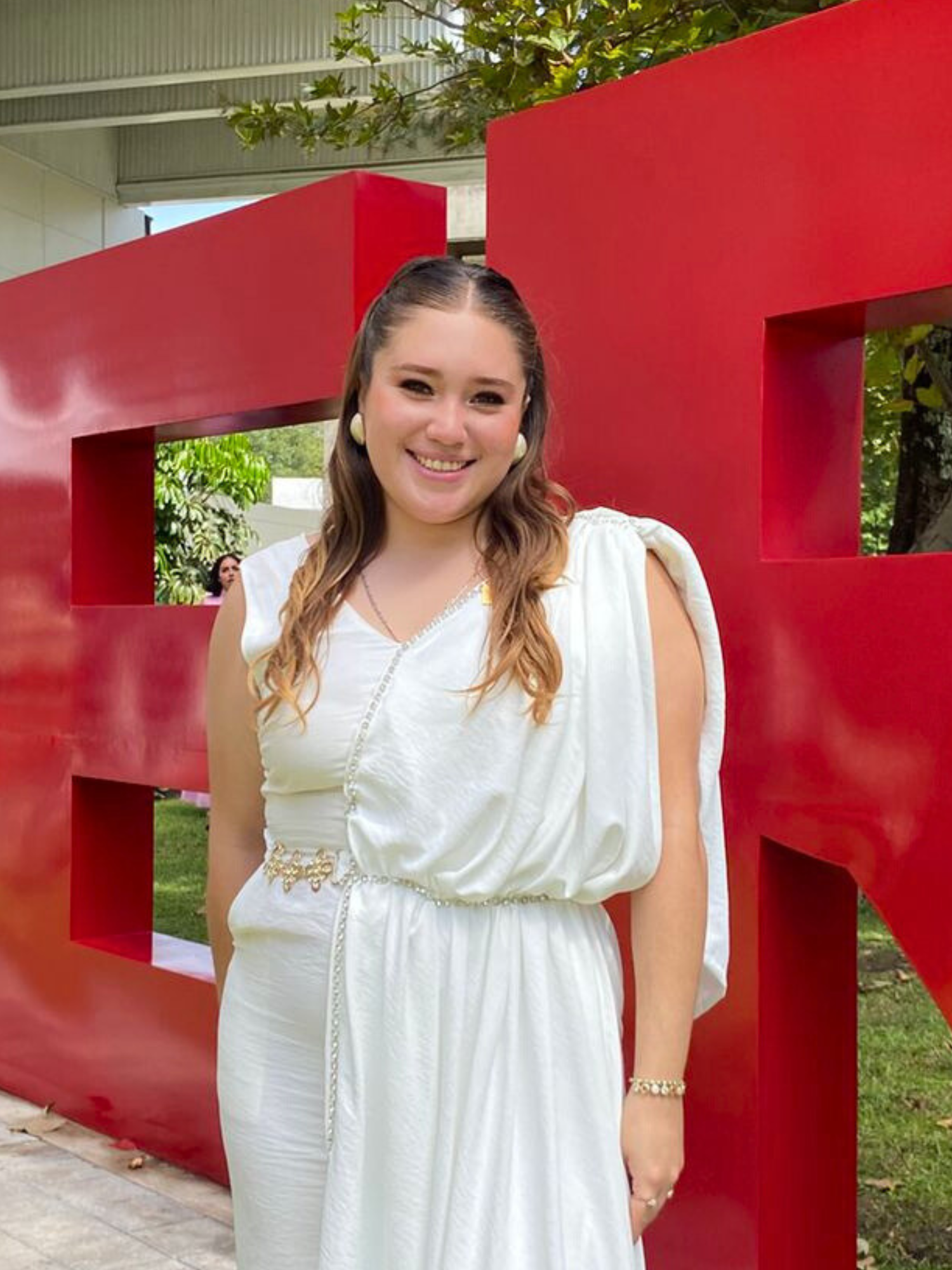 Joven mujer en vestido blanco, sonriendo, posando frente a una letra roja grande.
