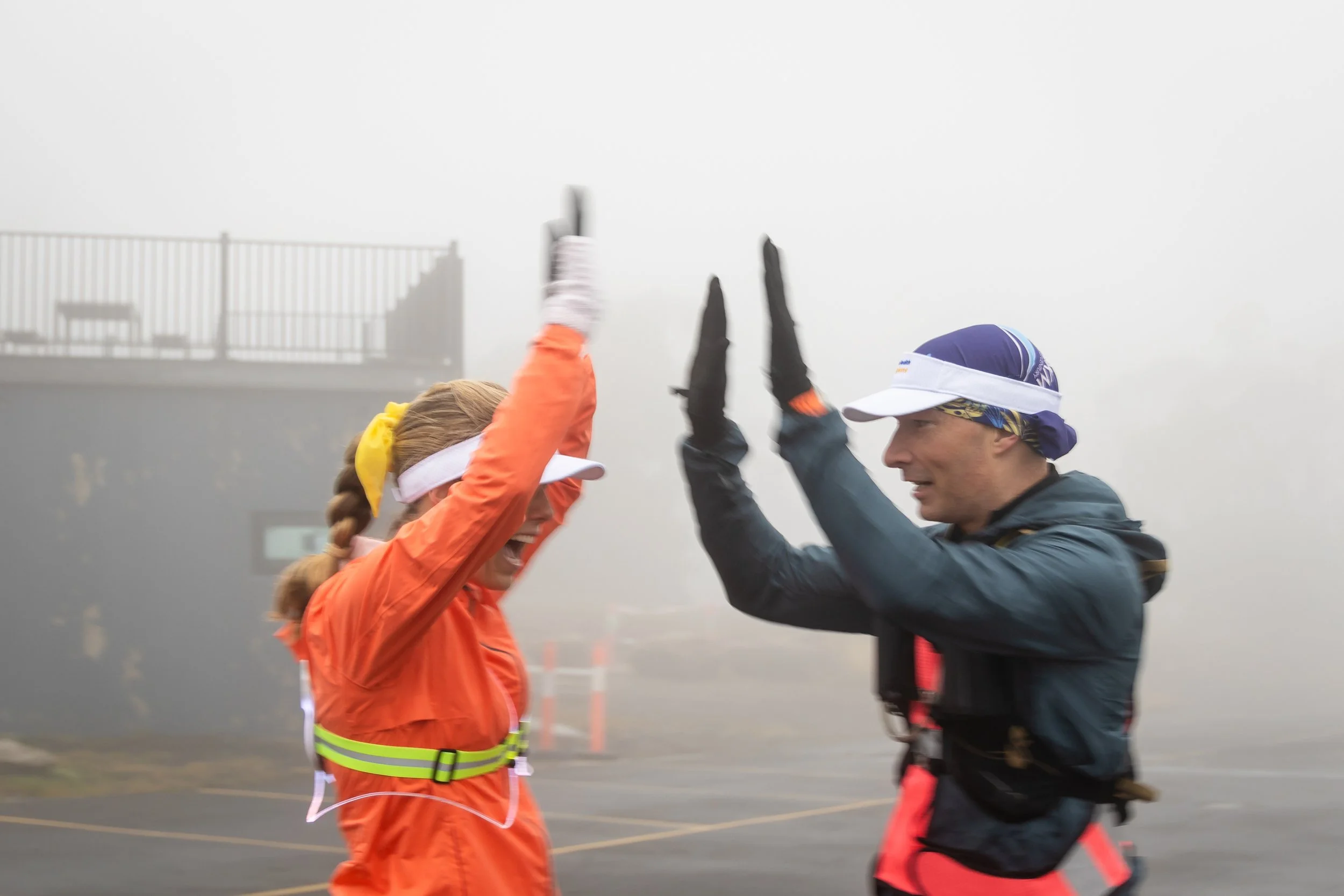 Two runners in athletic gear are celebrating with a high five in a foggy outdoor setting, possibly after a race.