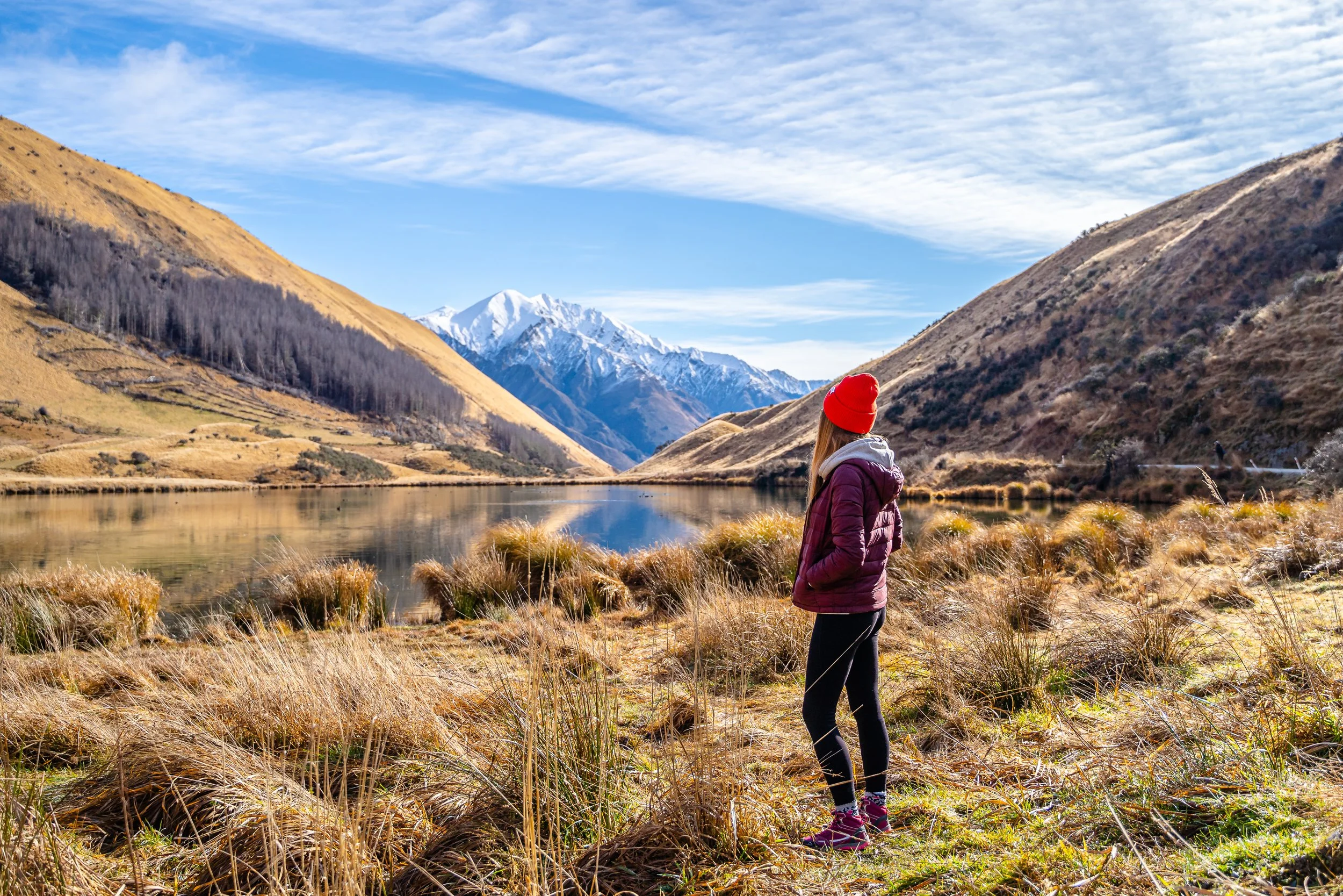 Person in red beanie and jacket standing by a lake in a mountainous landscape, with snow-capped peaks in the distance and dry grass in the foreground.