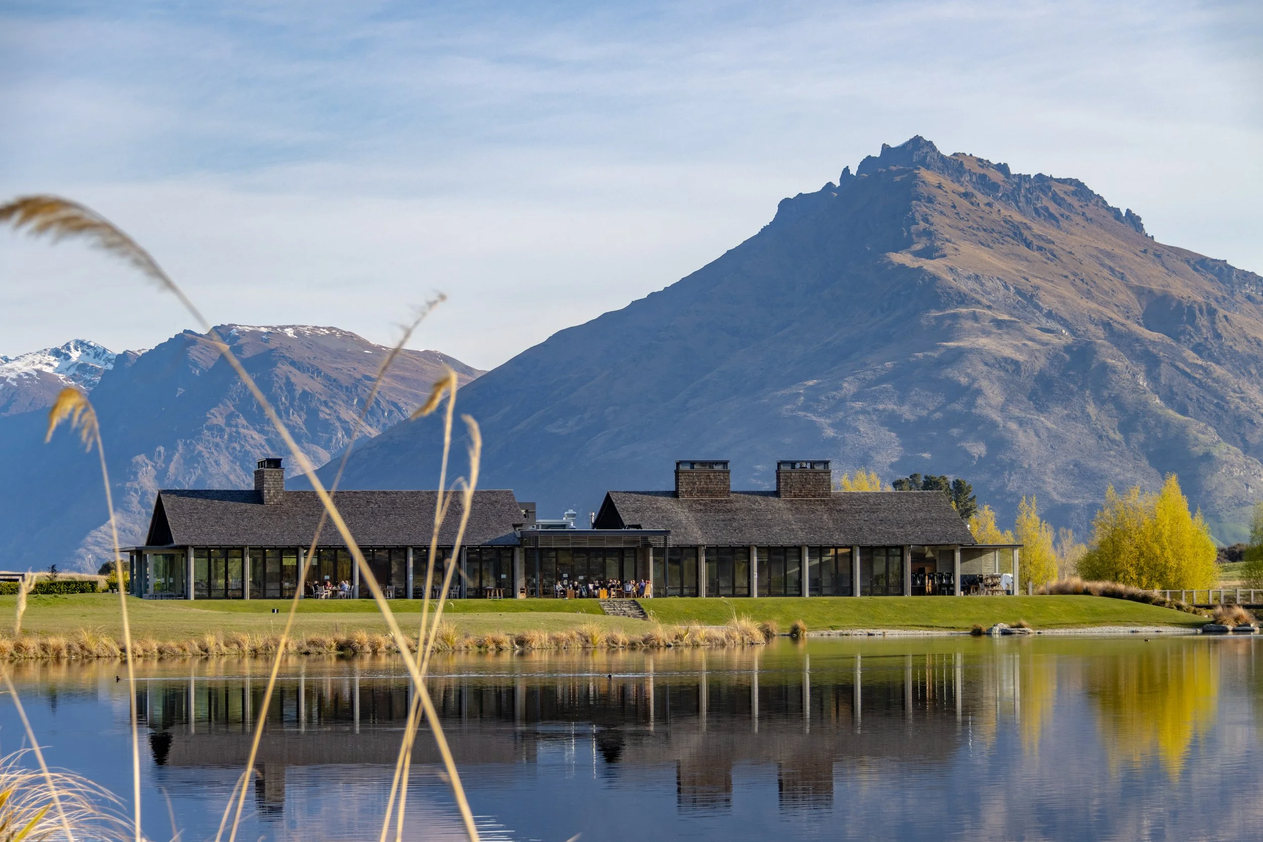 Modern building by serene lake with mountainous backdrop, surrounded by tall grass and trees.