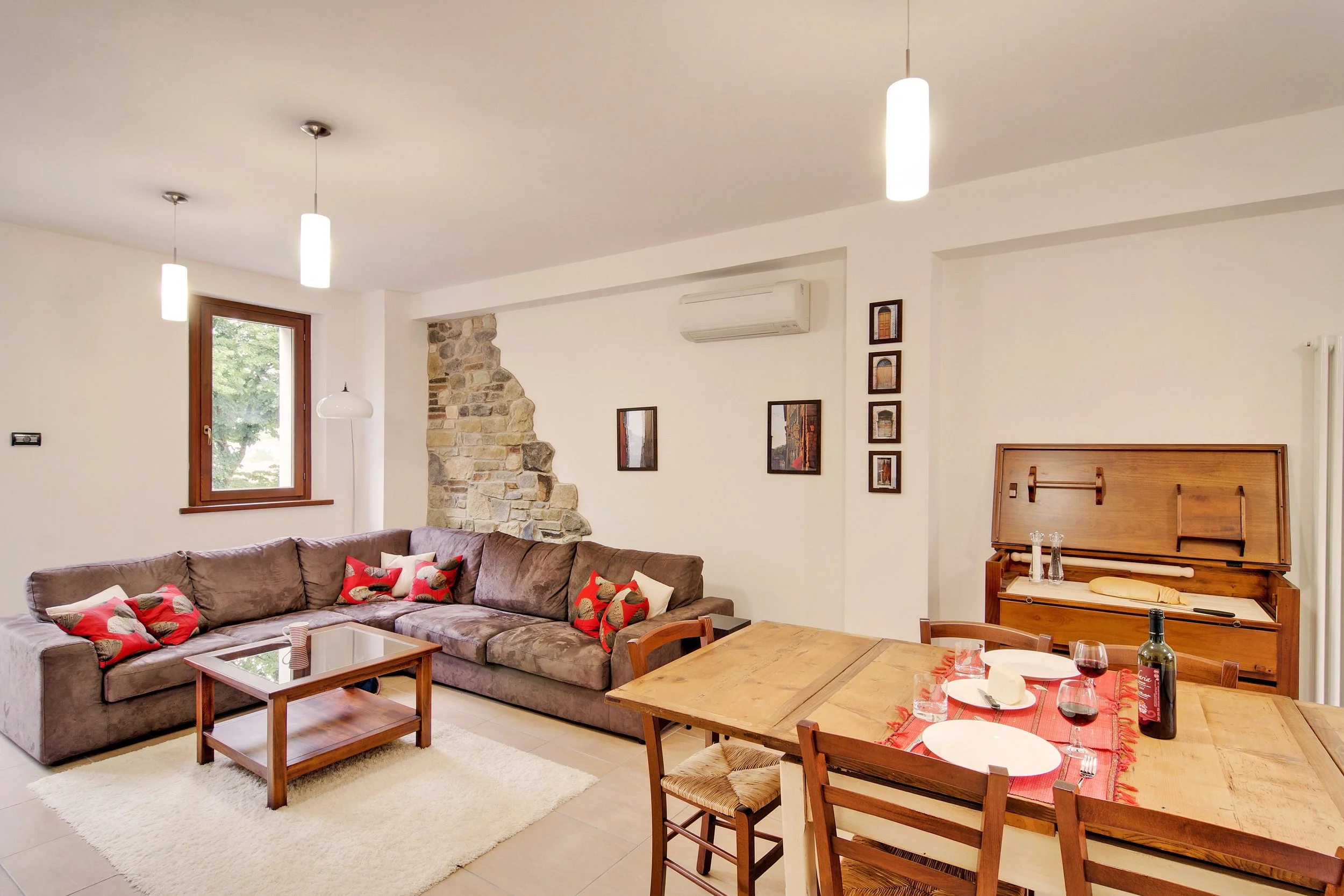 Cozy living room with a brown sectional sofa, red cushions, glass coffee table, wooden dining table, decorative stone wall, pendant lights, and small framed pictures.