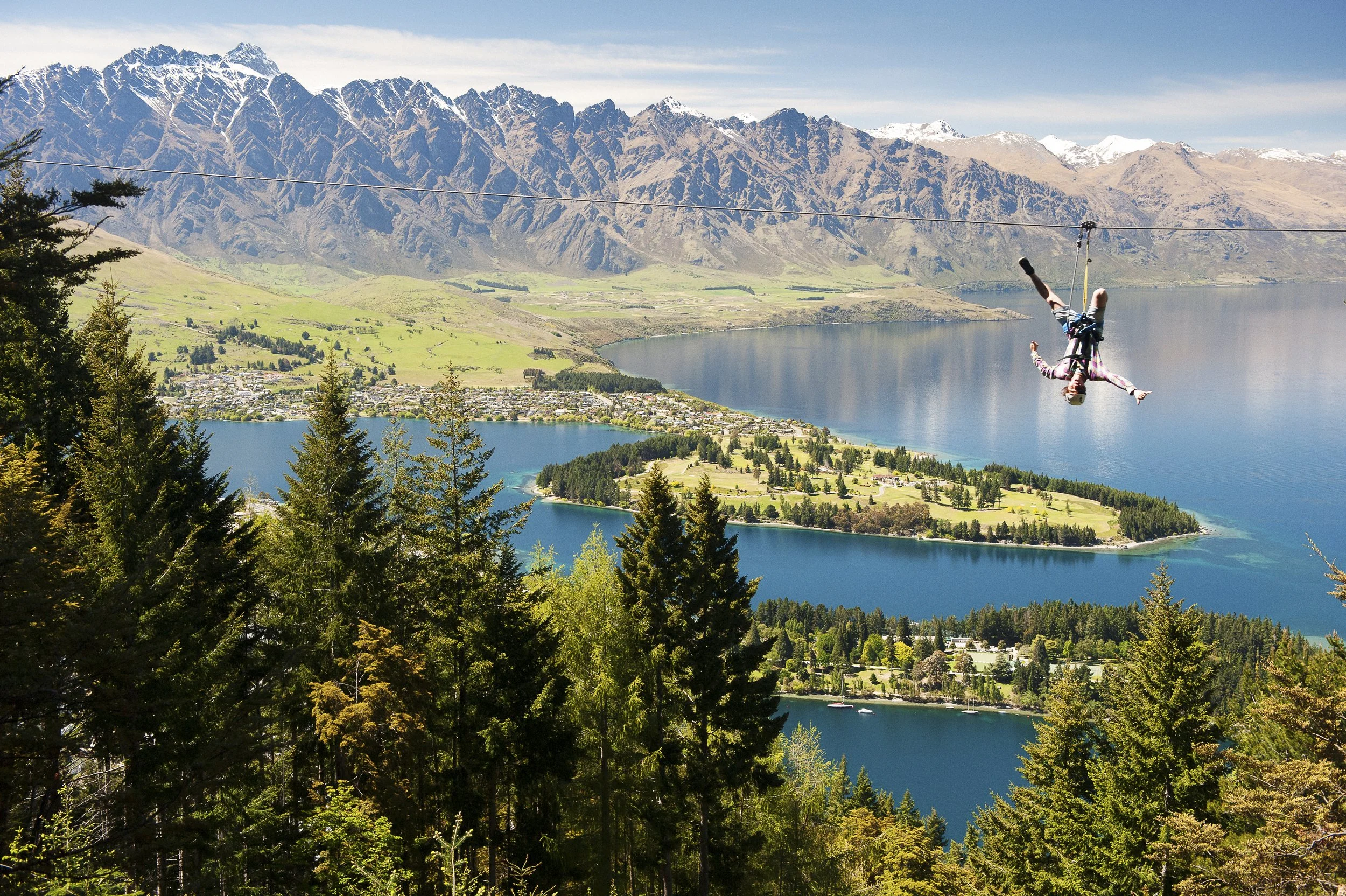 Person ziplining upside down over a forest with a lake and mountain range in the background.