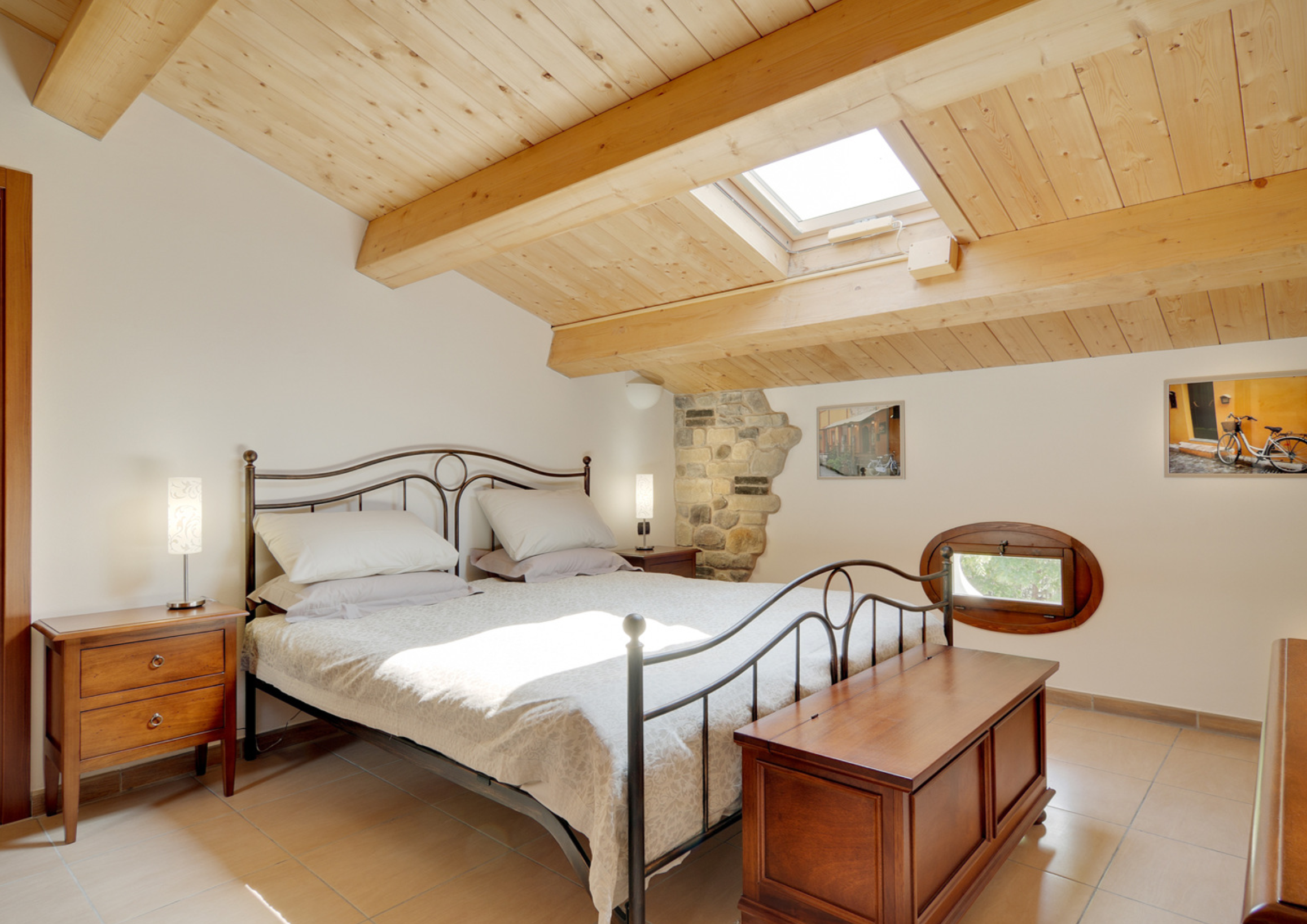 Cozy attic bedroom with wooden ceiling, skylight, metal bed frame, stone wall detail, and bedside tables.