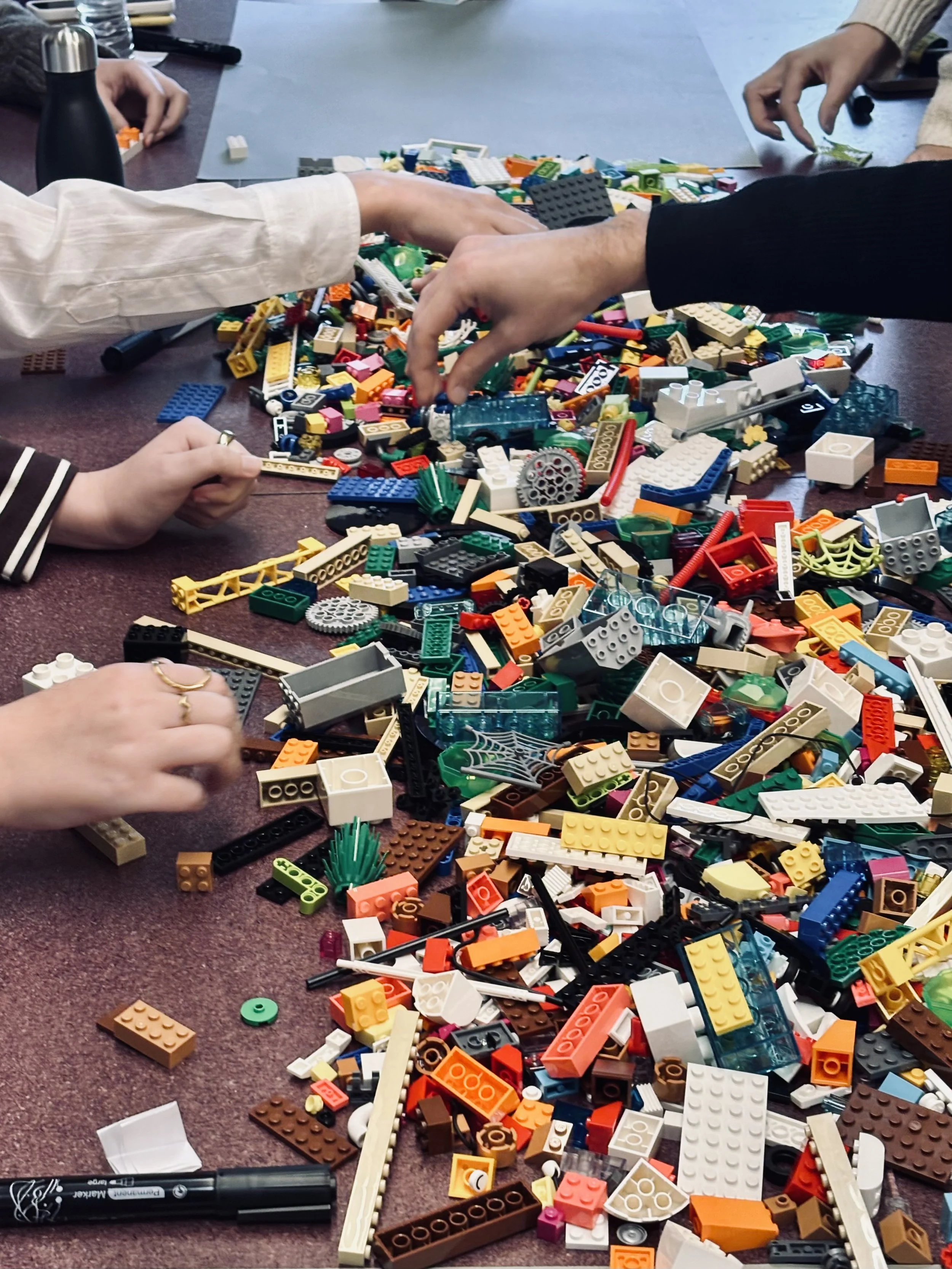 An assortment of LEGO bricks on a table as participants hands reach in to pick up bricks in the foreground and build models in the background