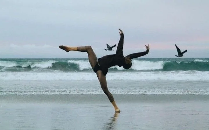 A woman dances with one leg raised in the air, gracefully arched back against a backdrop of the ocean with waves and two seagulls in flight