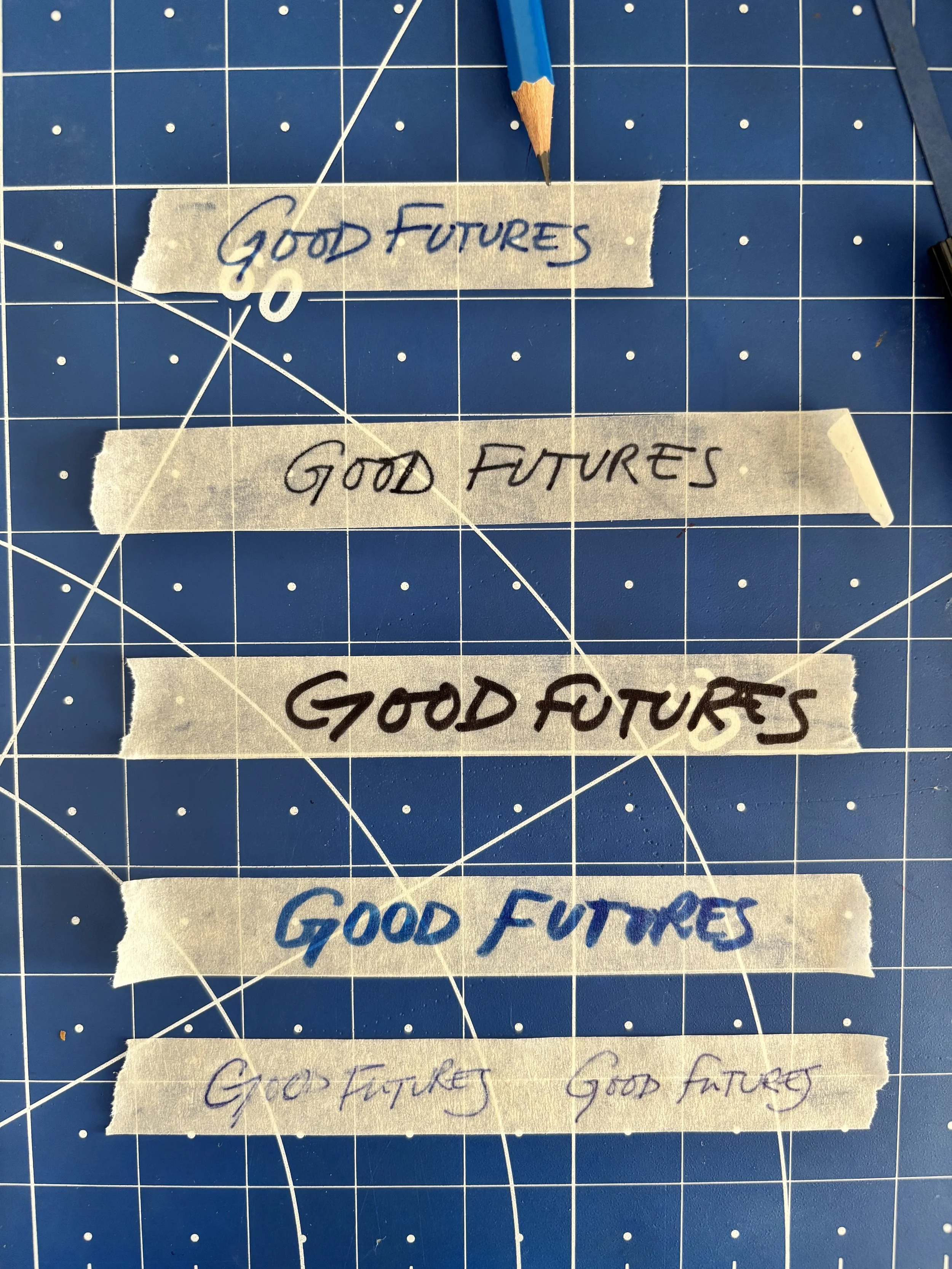 Three pieces of masking tape with handwritten text that says 'Good Futures' in blue, black, and purple ink respectively, placed on a blue cutting mat with white grid lines.