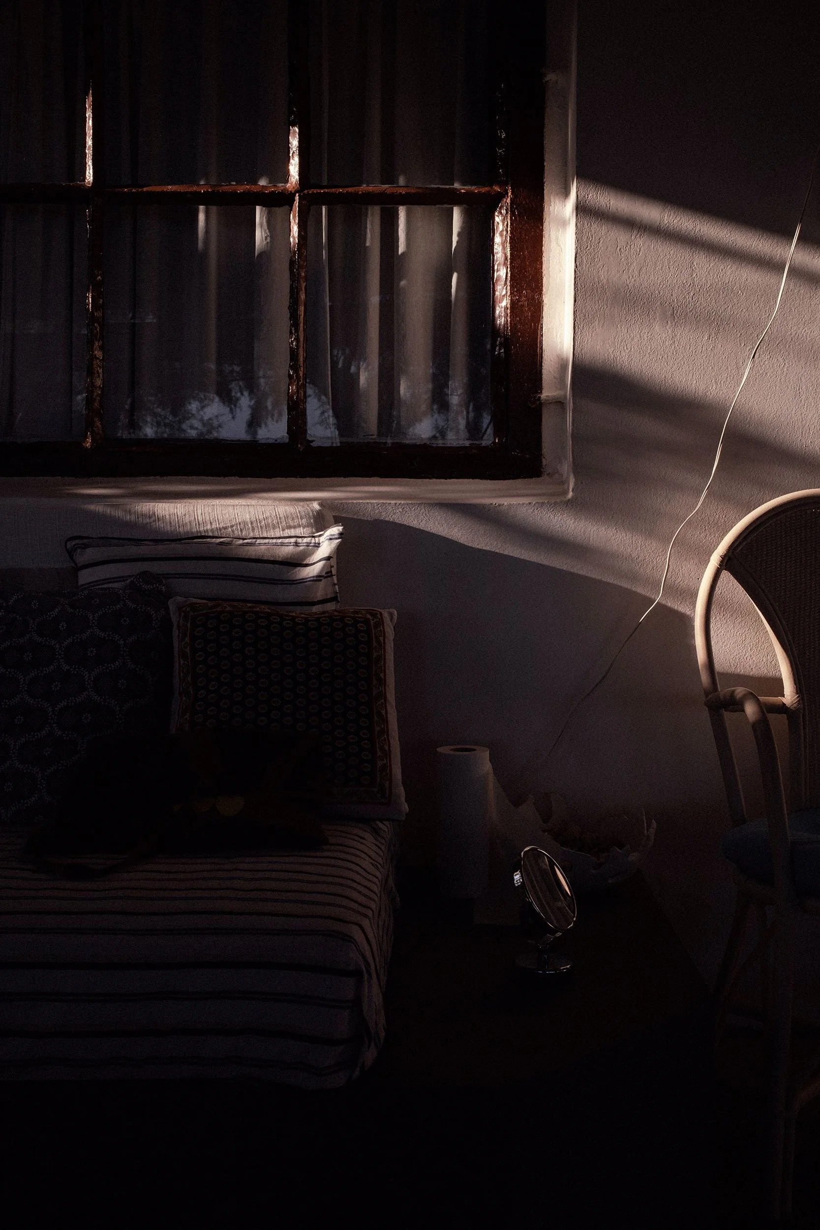 A dimly lit room with a bed, pillows, a window with bars, and a chair. Sunlight streams through the window casting shadows on the wall.