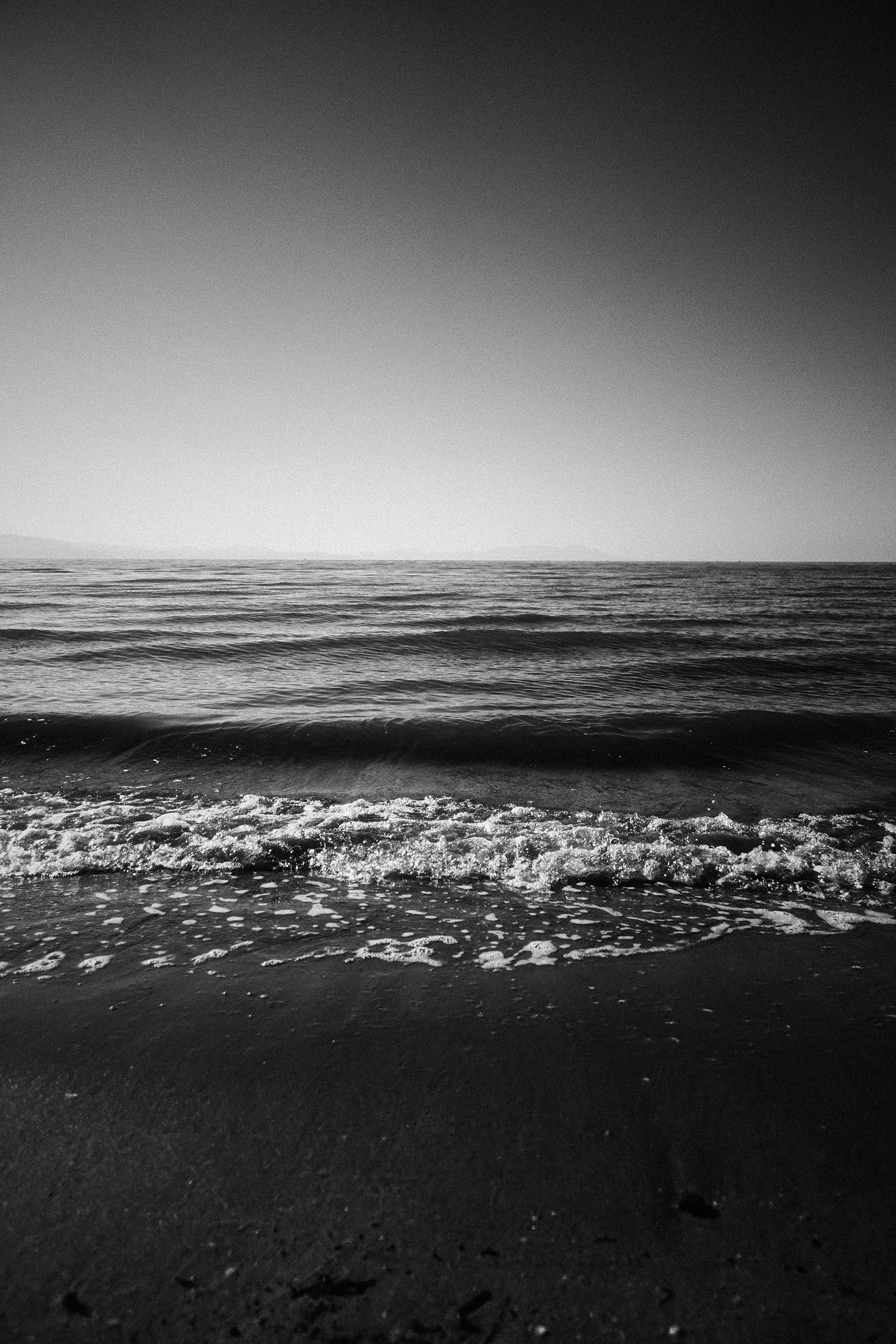 Black and white photo of ocean waves gently reaching the sandy beach, with a clear horizon and smooth sky.