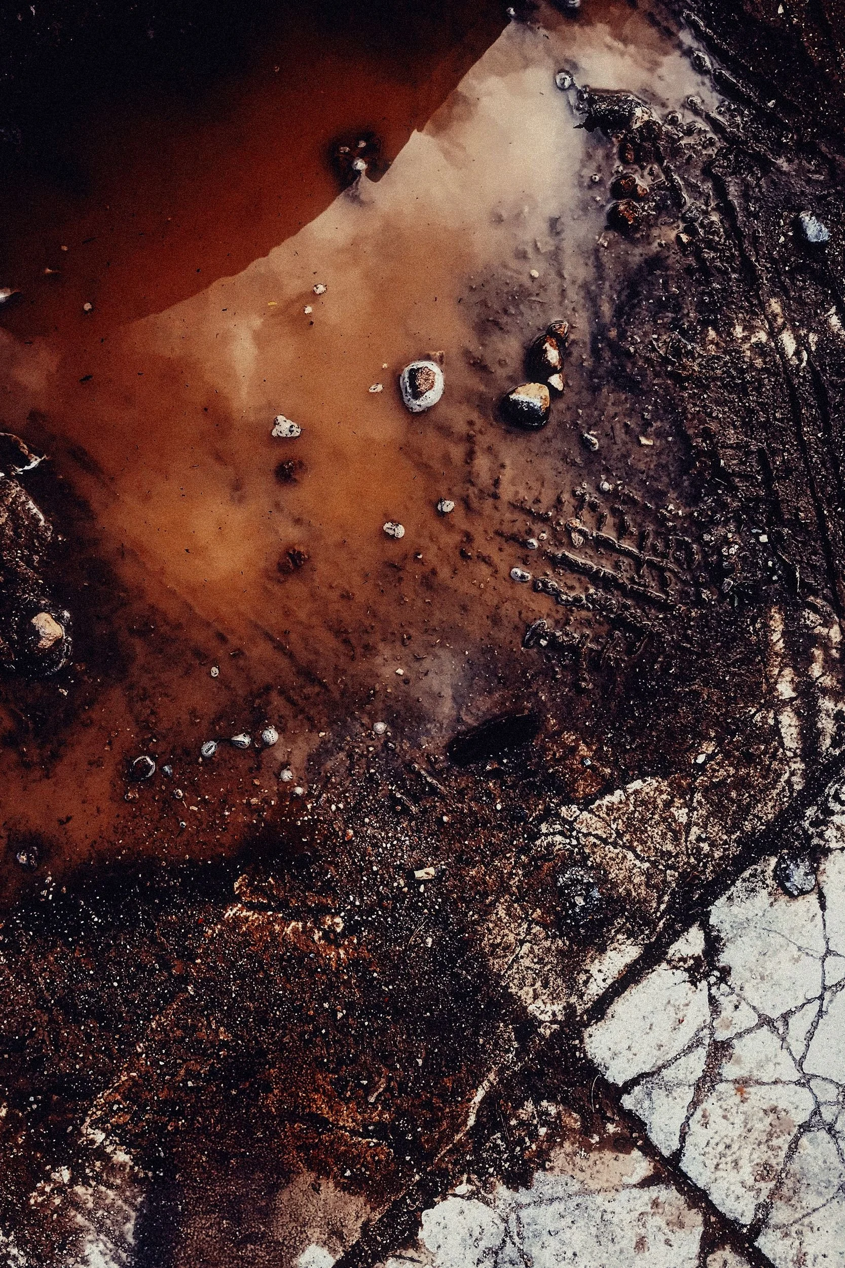 A close-up shot of a wet, cracked, and stained surface with small rocks and bubbles, possibly a concrete or asphalt ground.