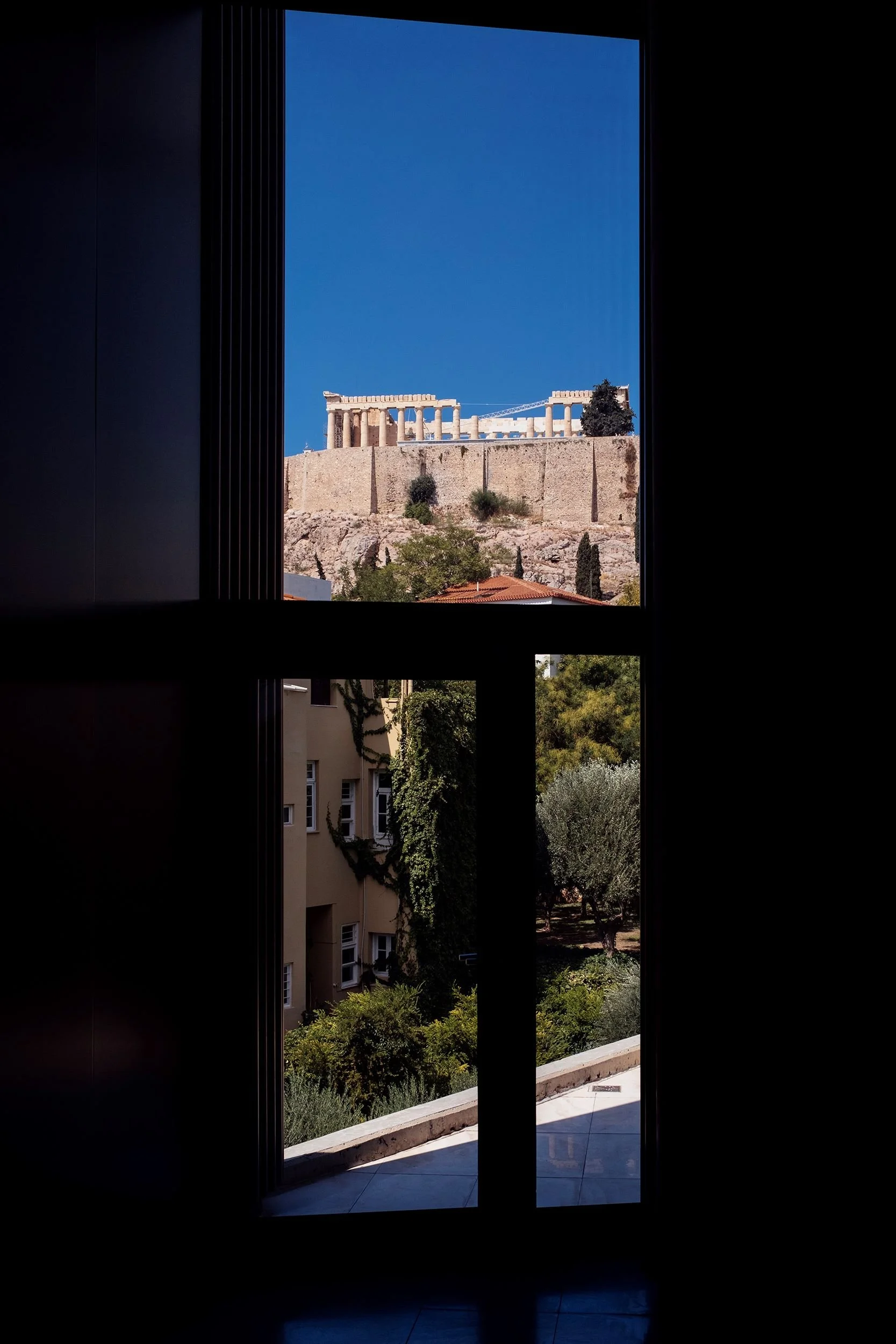 View of the Parthenon on the Acropolis in Athens, Greece, through a window with black framing, showing blue sky and surrounding trees.