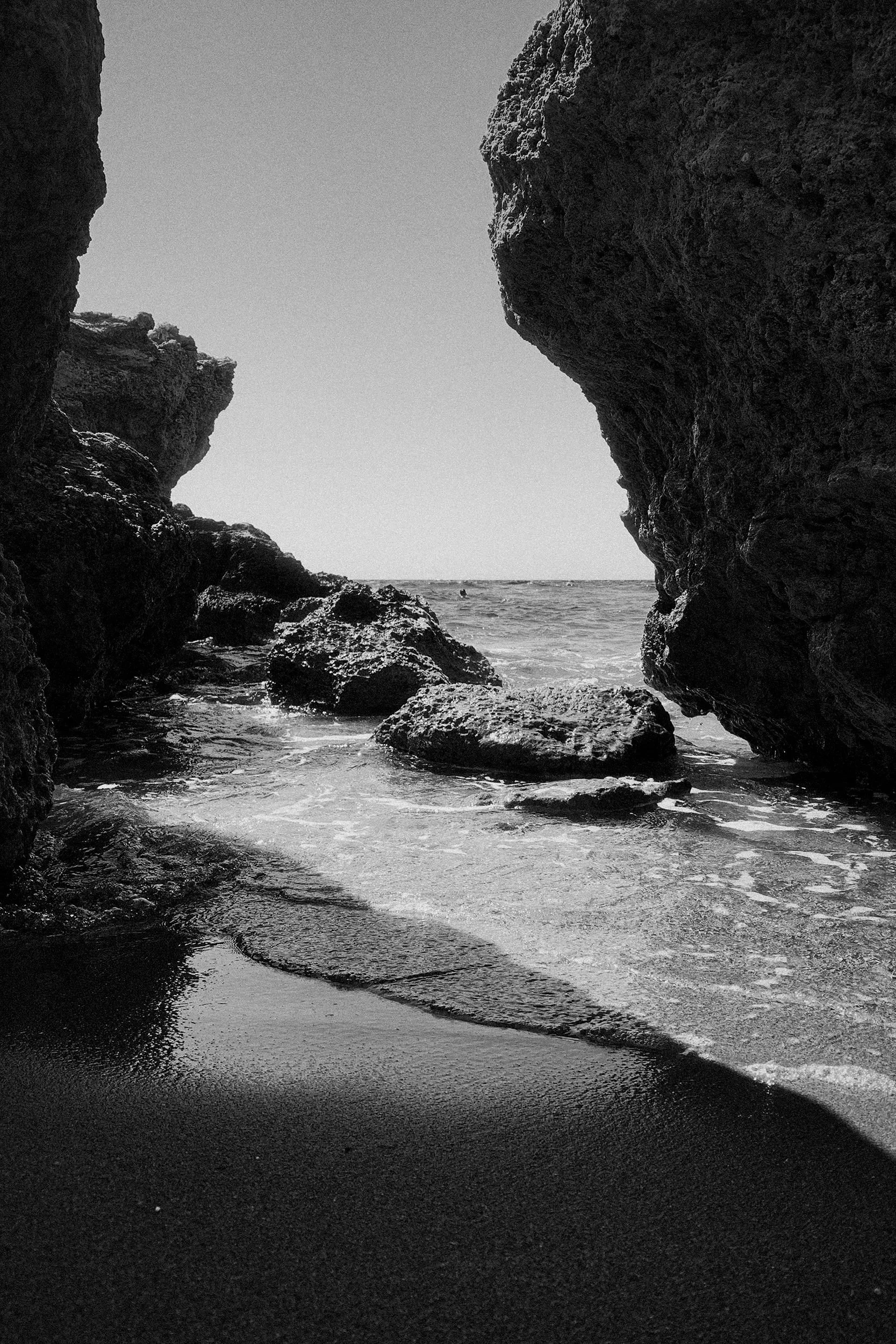 Black and white photo of a rocky coastal scene with a narrow opening between large rocks revealing the ocean and sky, with small waves on the sandy shore.