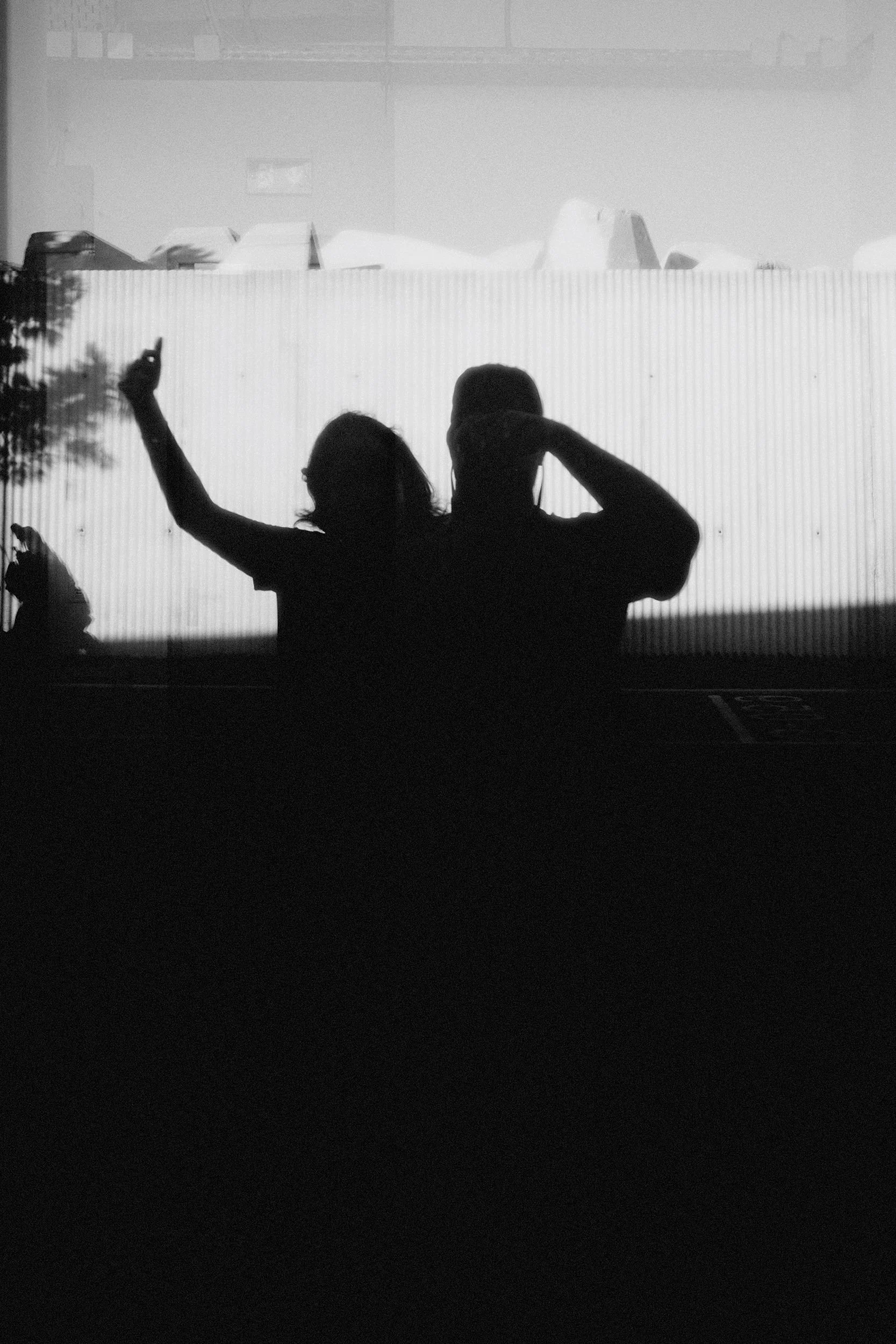 Silhouettes of two people sitting next to each other, with one person raising a hand and the other adjusting their hat, against a background of a wall or screen with some visible objects or structures.
