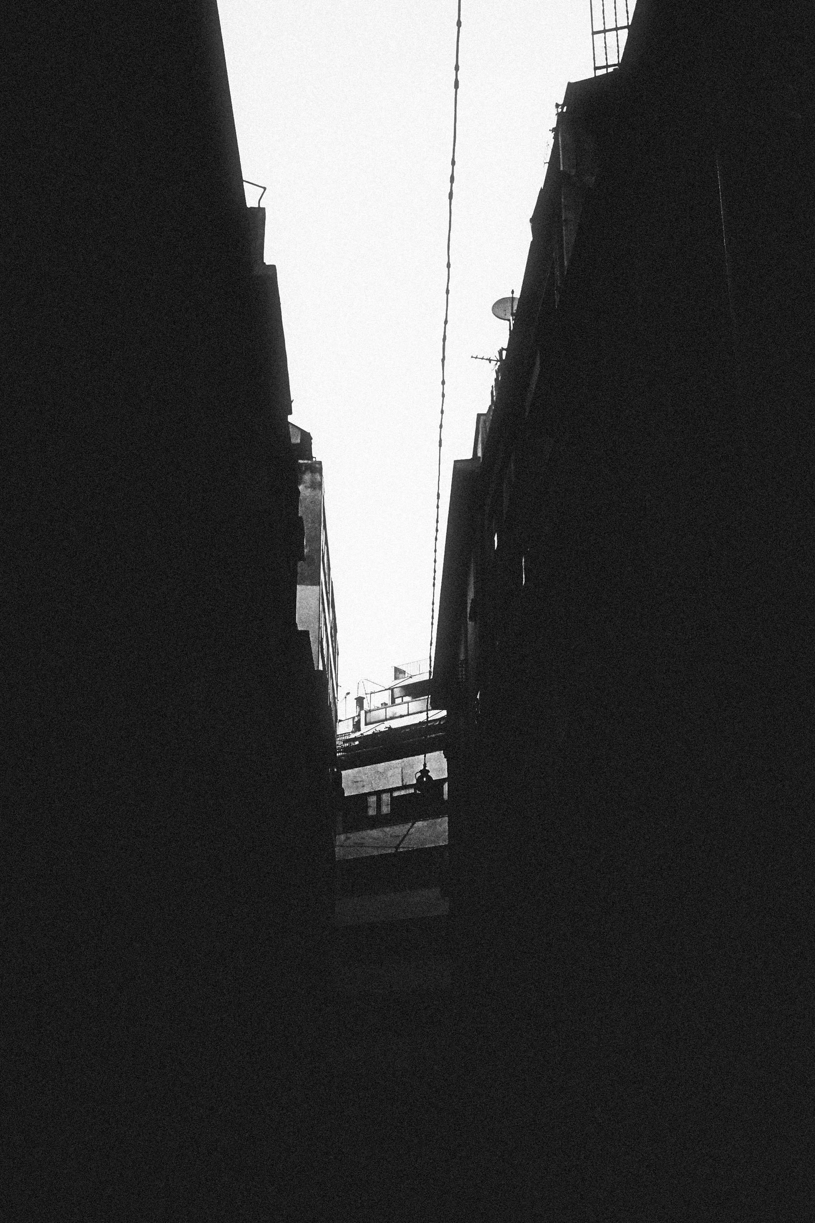 Black and white photo of a narrow alleyway between tall, dark buildings with a sky overhead and a bridge connecting the buildings in the background.