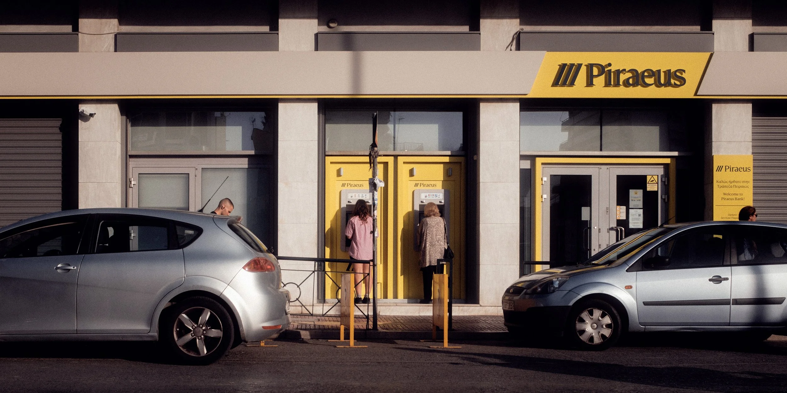 Color photograph of people using ATMs outside a Piraeus Bank in Athens, Greece. Framed by parked cars and afternoon light, the image captures an authentic urban moment of everyday city life.
Keywords: Athens street photography, Piraeus Bank, Greek ur