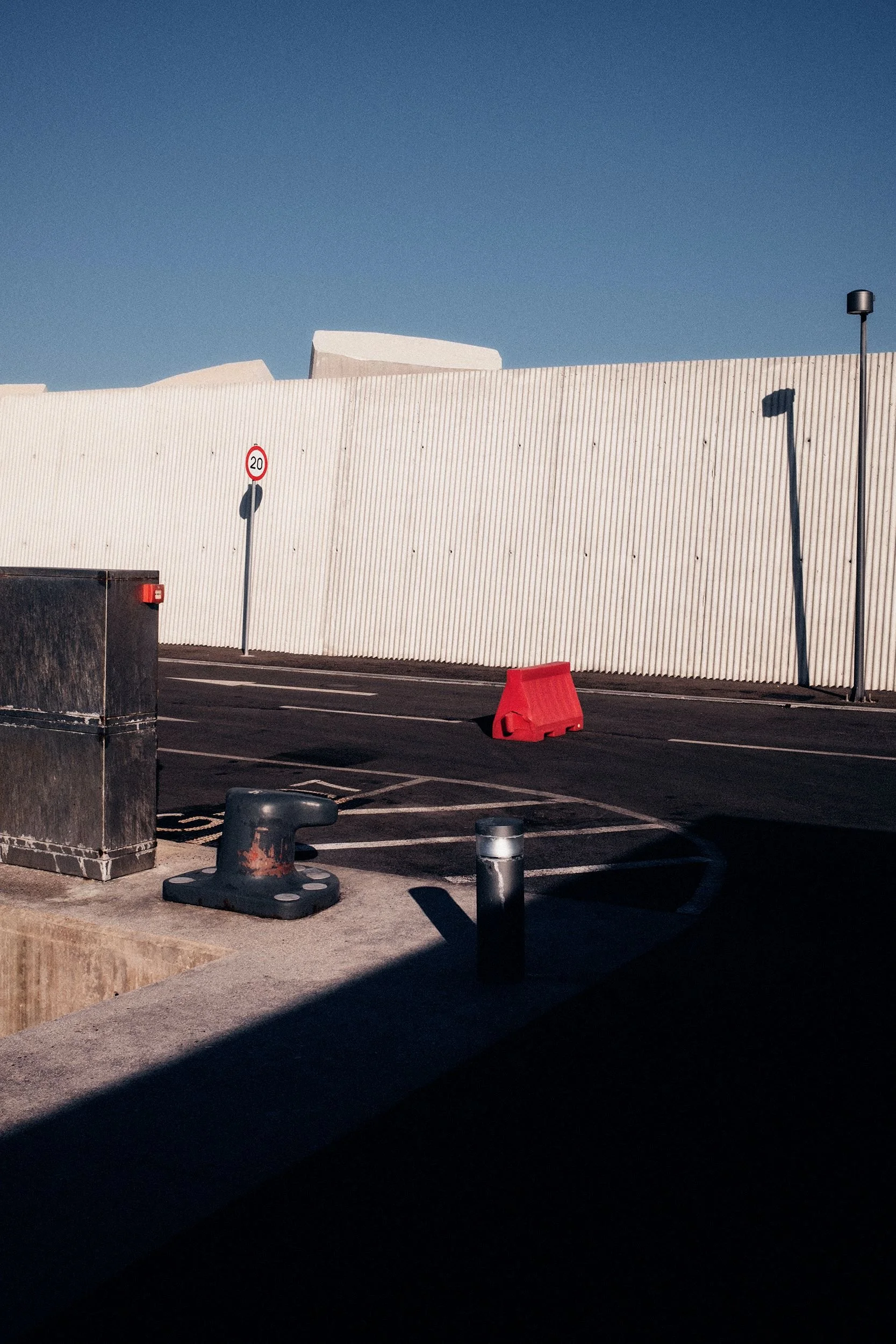 Empty parking lot with a red traffic barrier, a 20 mph speed limit sign, a black trash bin, and shadows cast by two streetlights against a white corrugated wall under a clear blue sky.