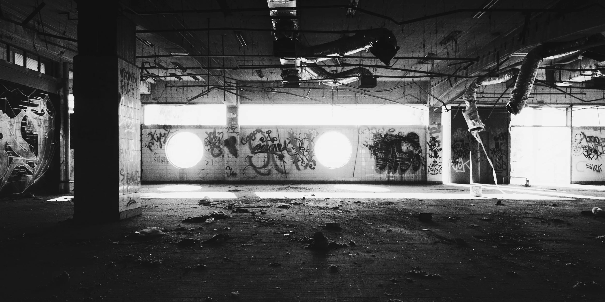 Black and white photograph of an abandoned industrial warehouse with graffiti-covered walls and circular windows. The decaying ceiling pipes, broken floor debris, and natural light filtering through create a raw urban atmosphere — ideal for editorial