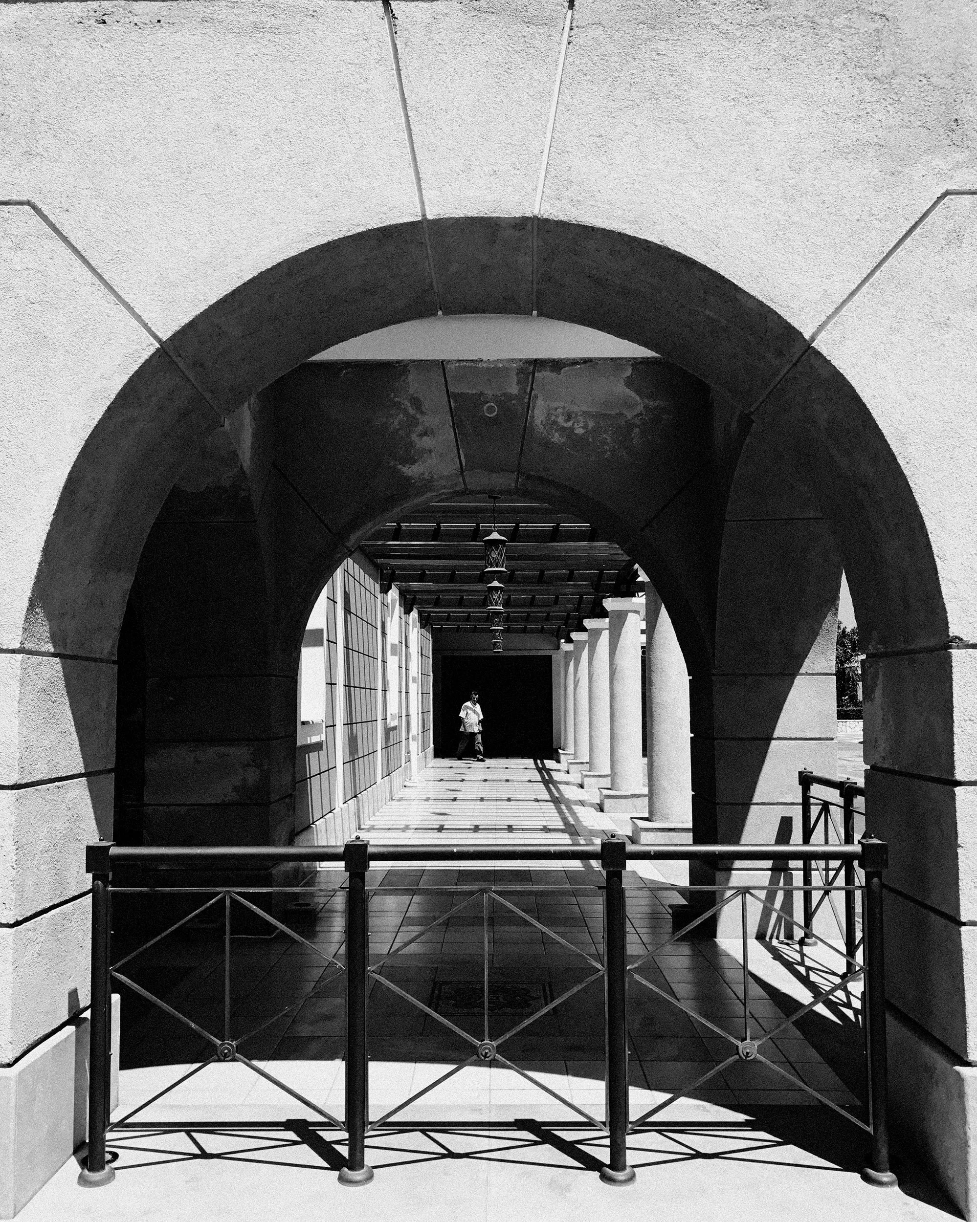 Black and white photograph of a corridor with arches and columns, shadow cast across the walkway, a person walking in the distance.