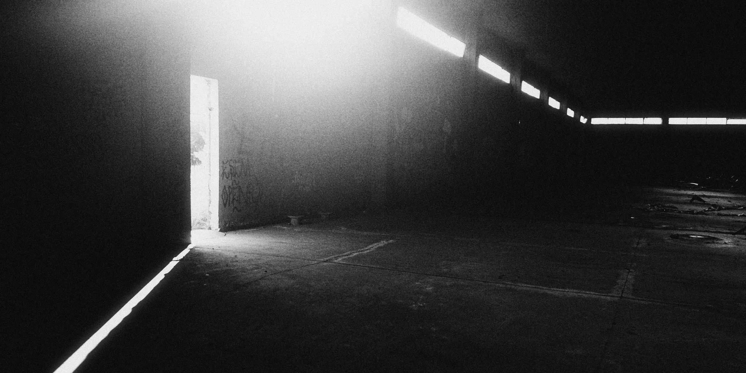 Fine art black and white image of sunlight piercing through a doorway in an empty, dark industrial hall. The contrast of shadow and light creates a haunting, cinematic mood.
Keywords: black and white photography, light leak, abandoned space, cinemati