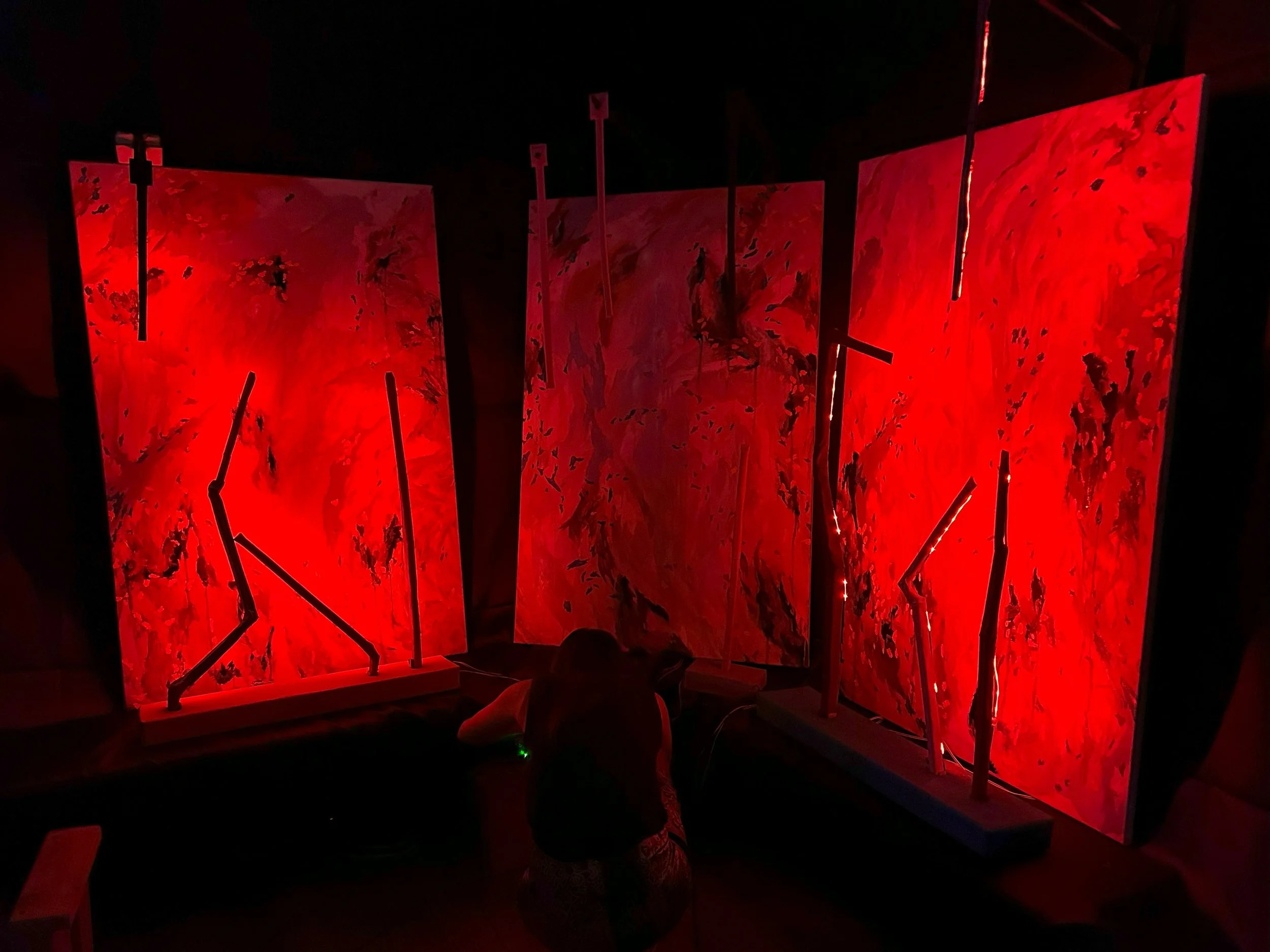 Three large red abstract paintings with black metal brackets holding them upright in a dark room, illuminated by red lighting, with a person crouching in front.