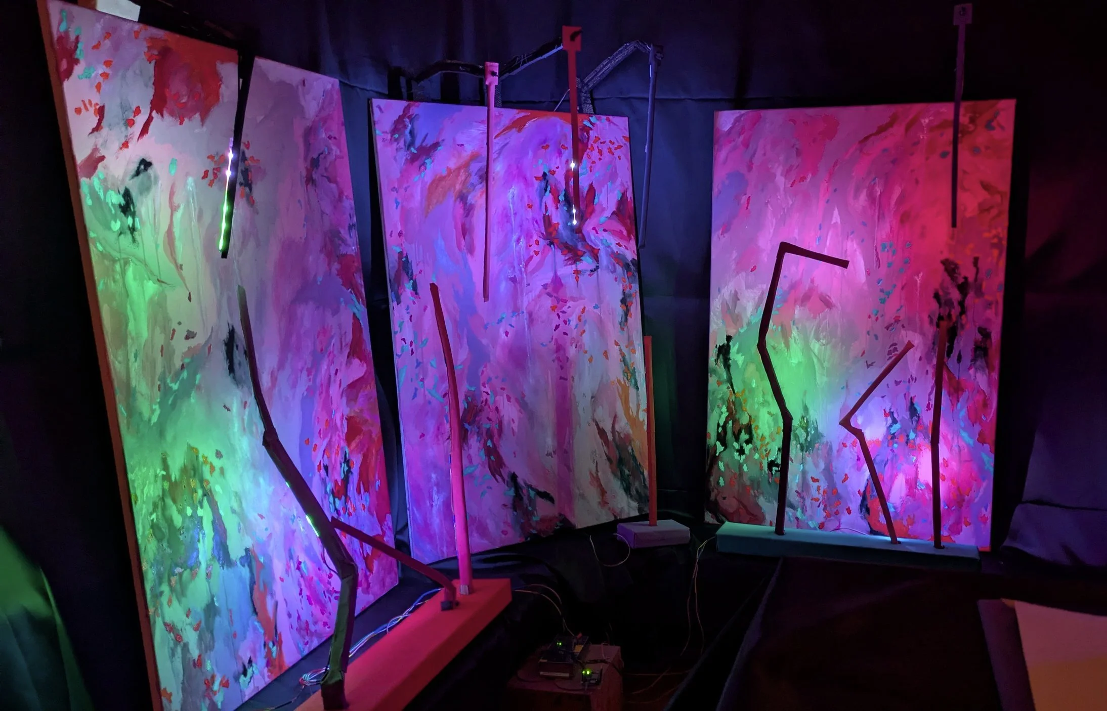 Three abstract, colorful paintings illuminated by neon lights in a dark room, with visible electronic equipment on the floor.