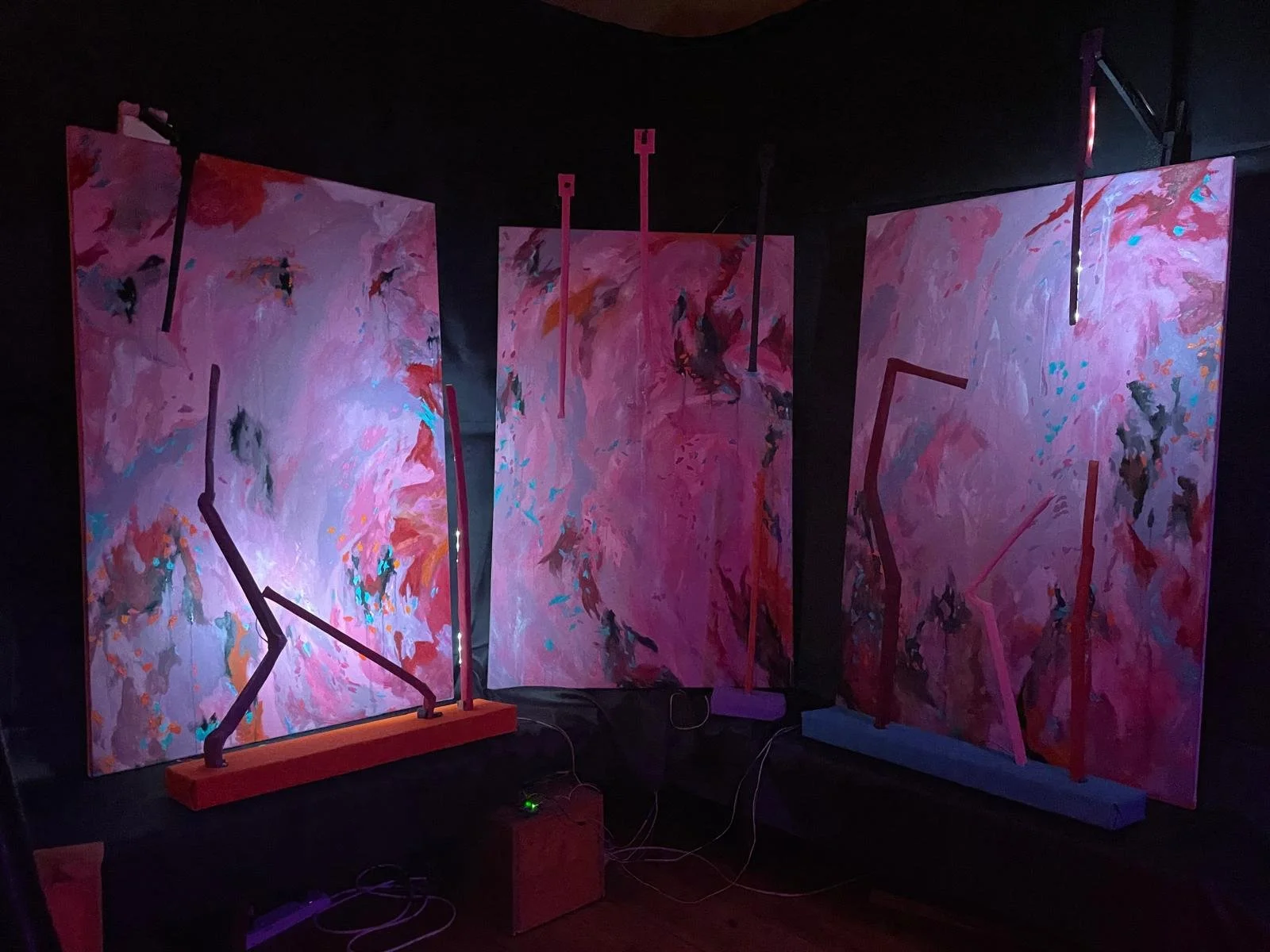 Three abstract paintings with pink, red, purple, and black brushstrokes, each illuminated by a small black light fixture, set against a dark background.