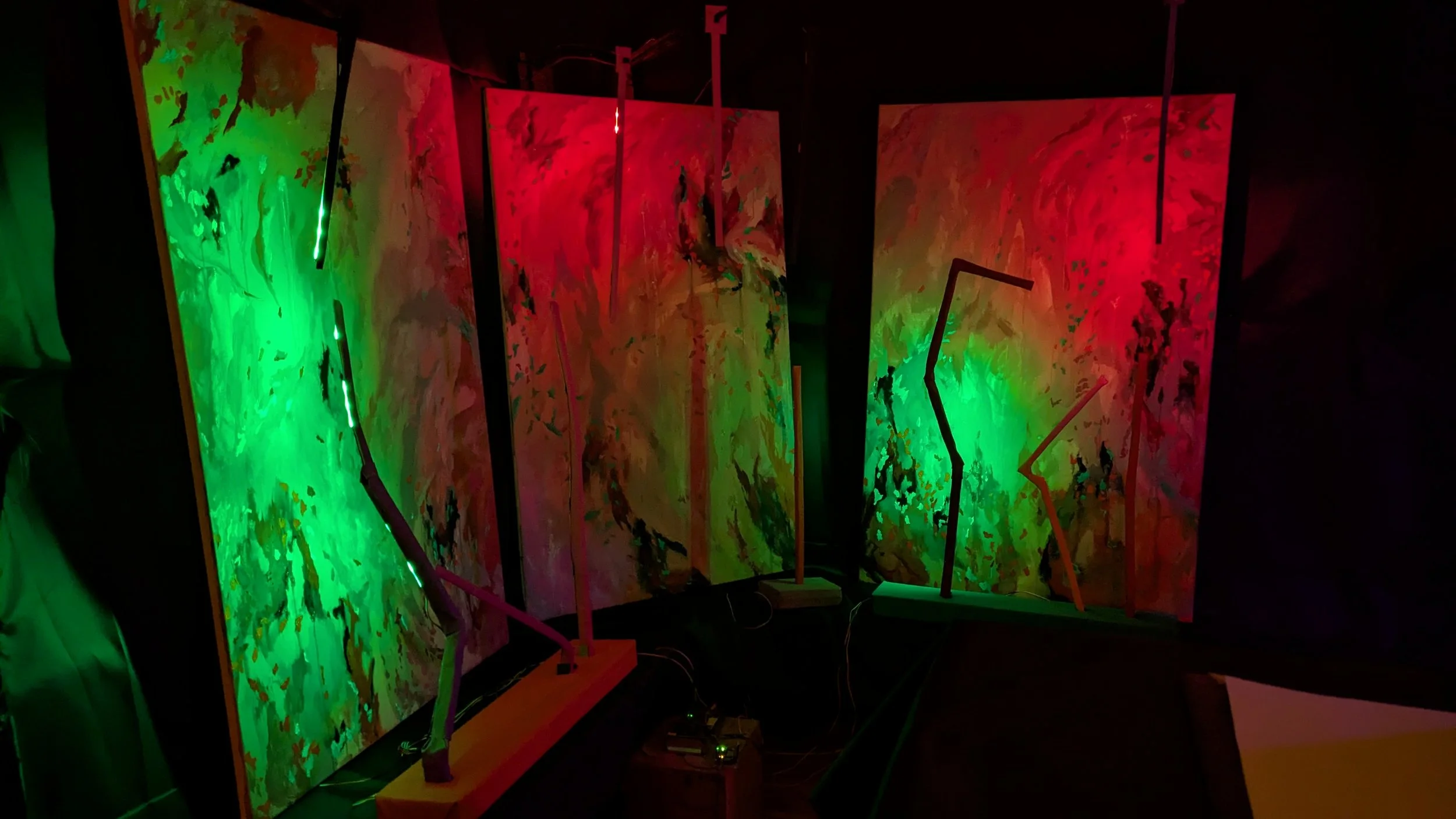 Three abstract paintings with vibrant green and red colors are displayed on a dark wall, each illuminated by LED lights attached to bent rods on wooden bases.