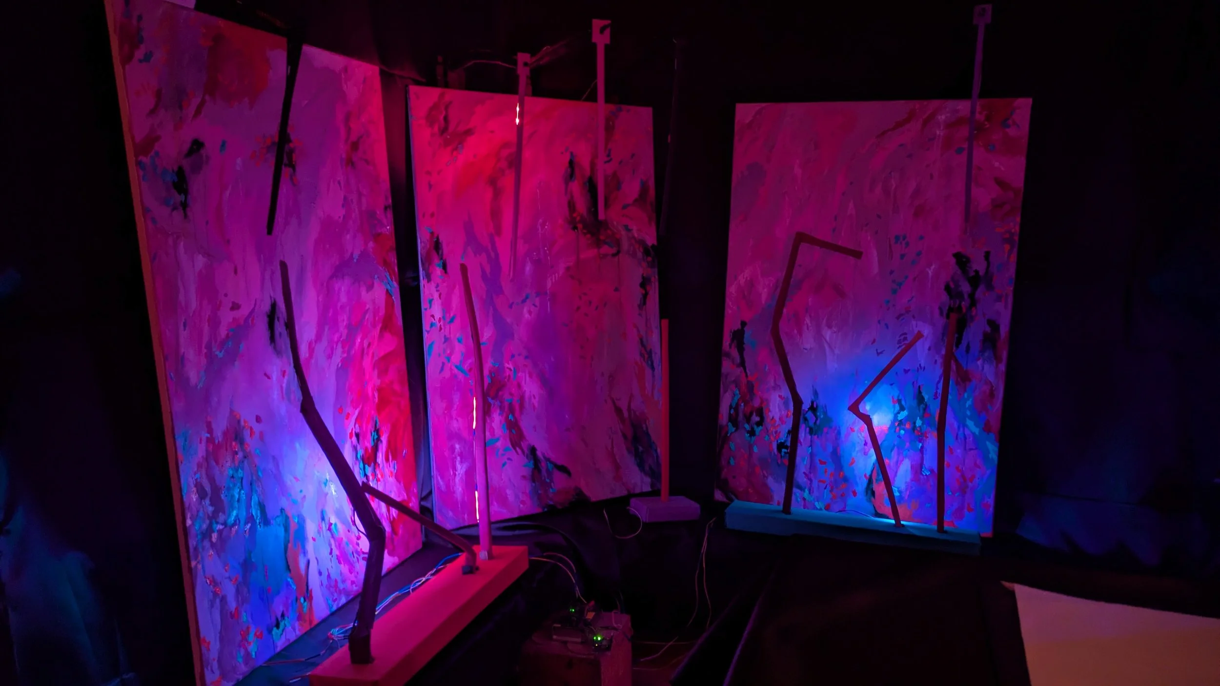 Abstract colorful paintings with neon lighting on display at an art exhibit, featuring shades of pink, purple, blue, and black.